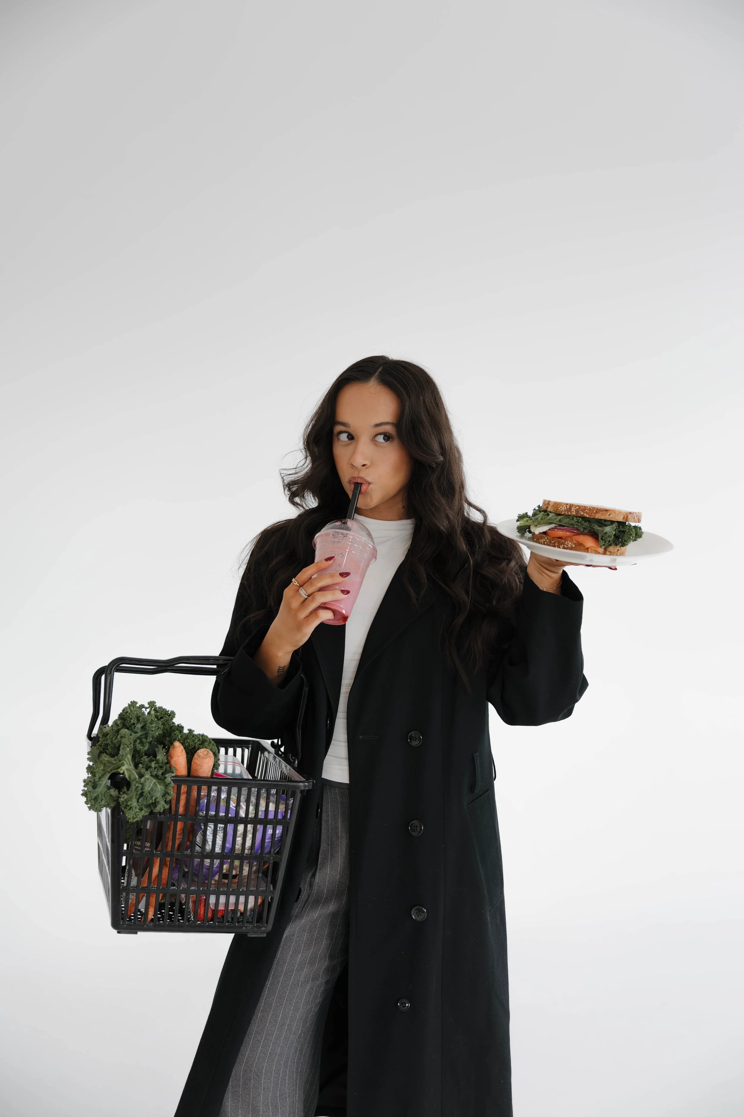 A woman with long dark curly hair, wearing a black long coat and gray pinstripe pants, holding a shopping basket filled with groceries in one hand and a plate with a sandwich in the other hand, sipping a pink beverage through a black straw.