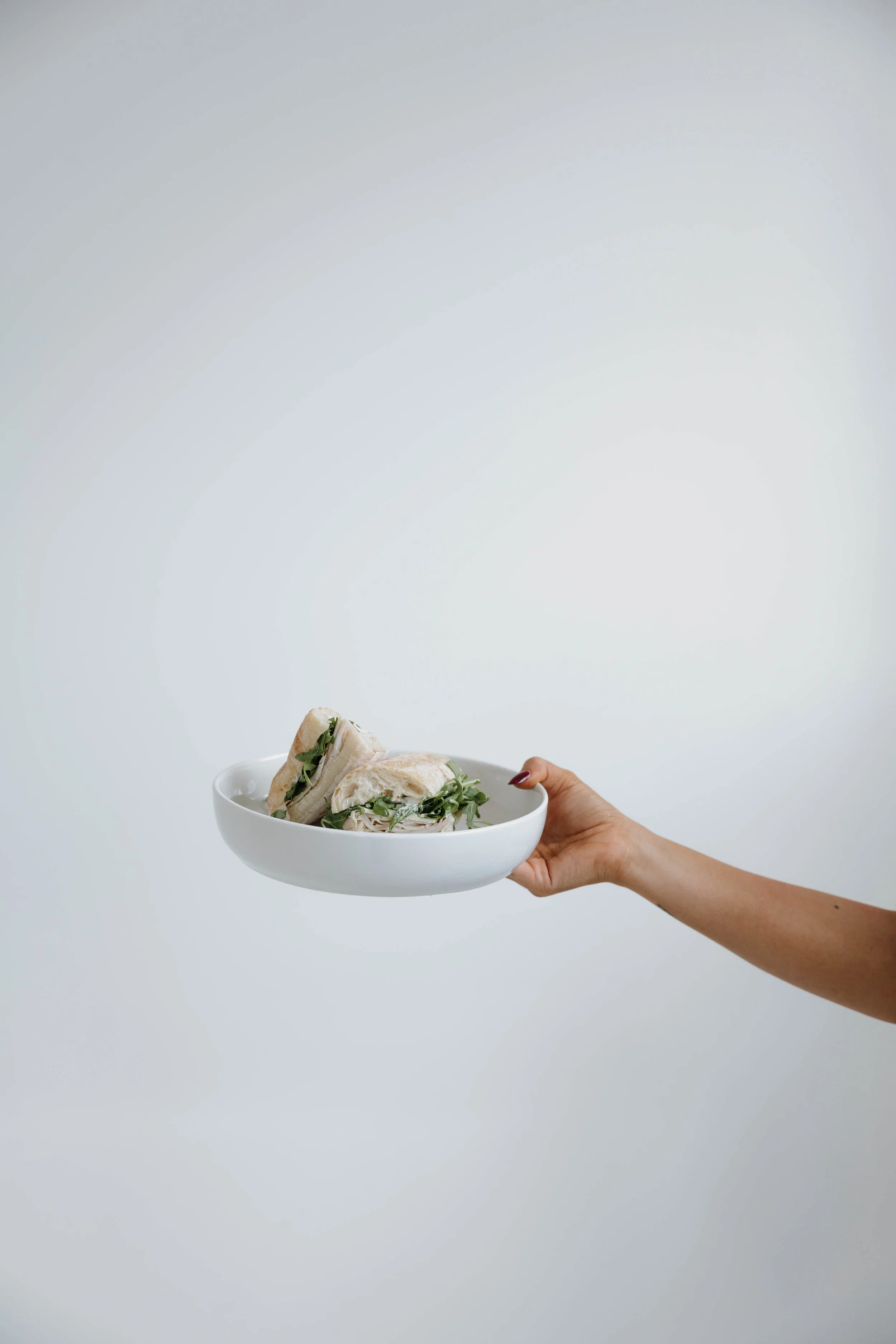 A hand holding a white bowl with sandwich halves filled with greens and other ingredients against a plain background.