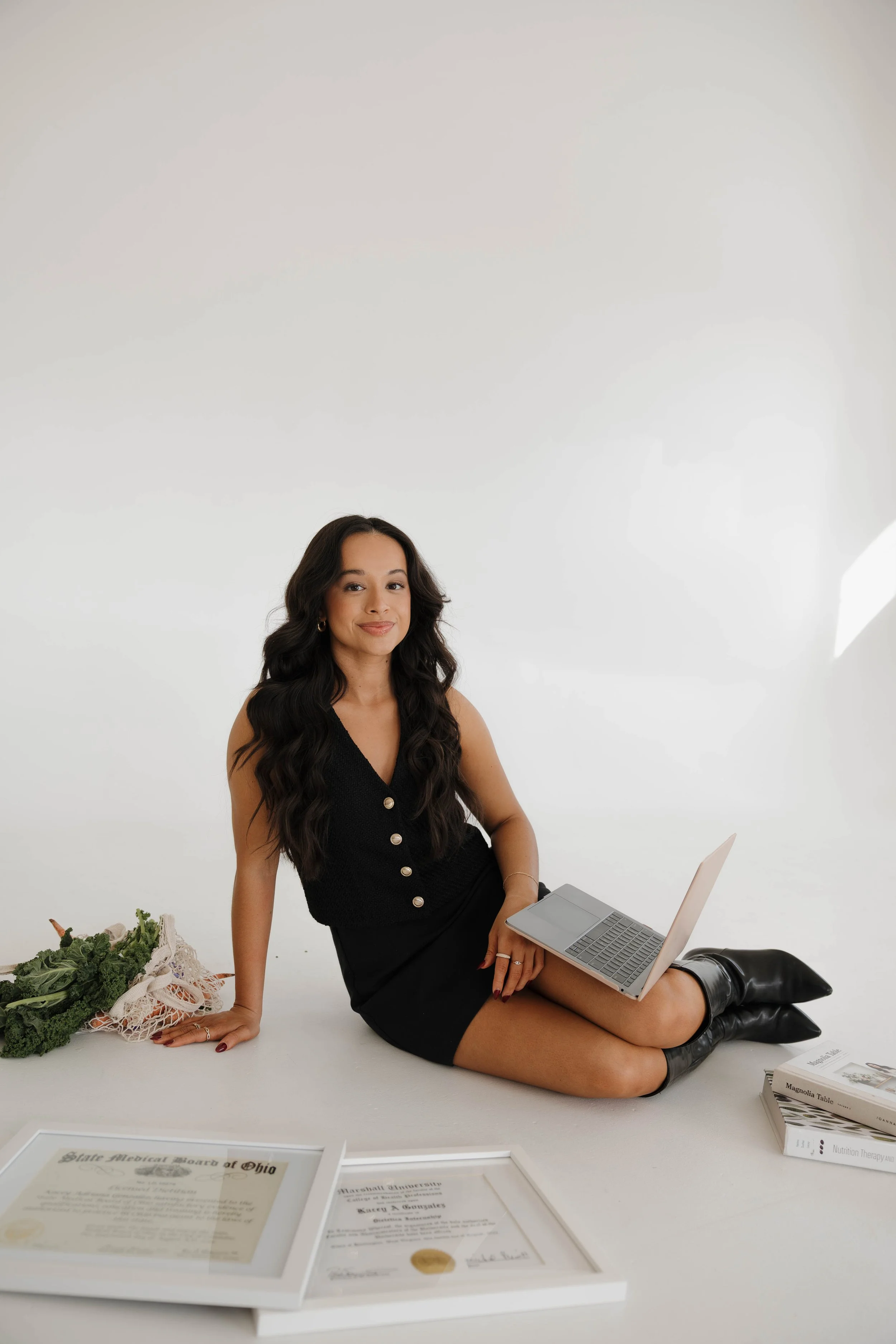 A woman with long dark hair, sitting on the floor with a laptop on her lap, smiling at the camera, surrounded by framed certificates, vegetables, and books in a white studio setting.
