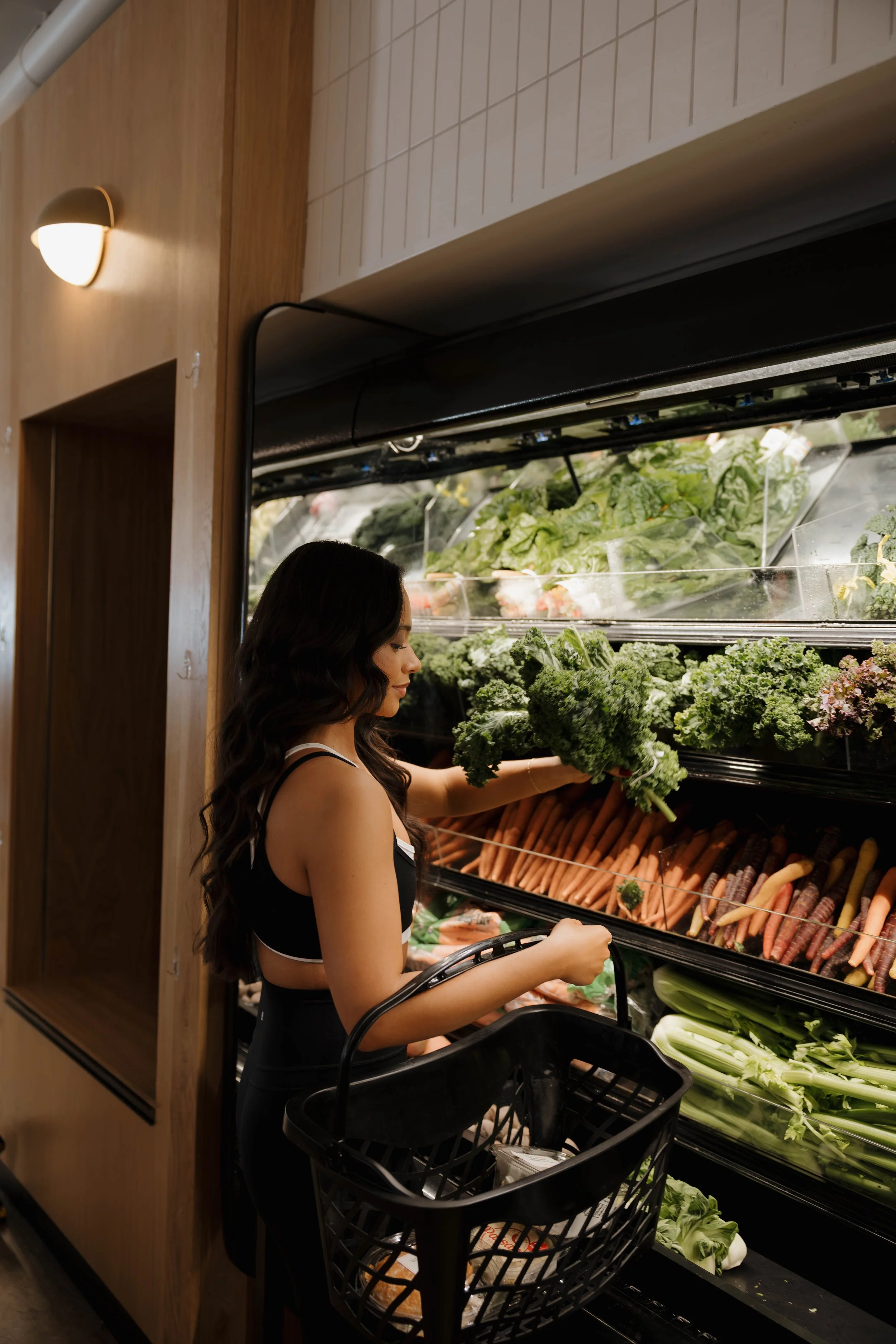 A woman shopping for vegetables at a grocery store, selecting kale from the produce section with a shopping basket in hand.