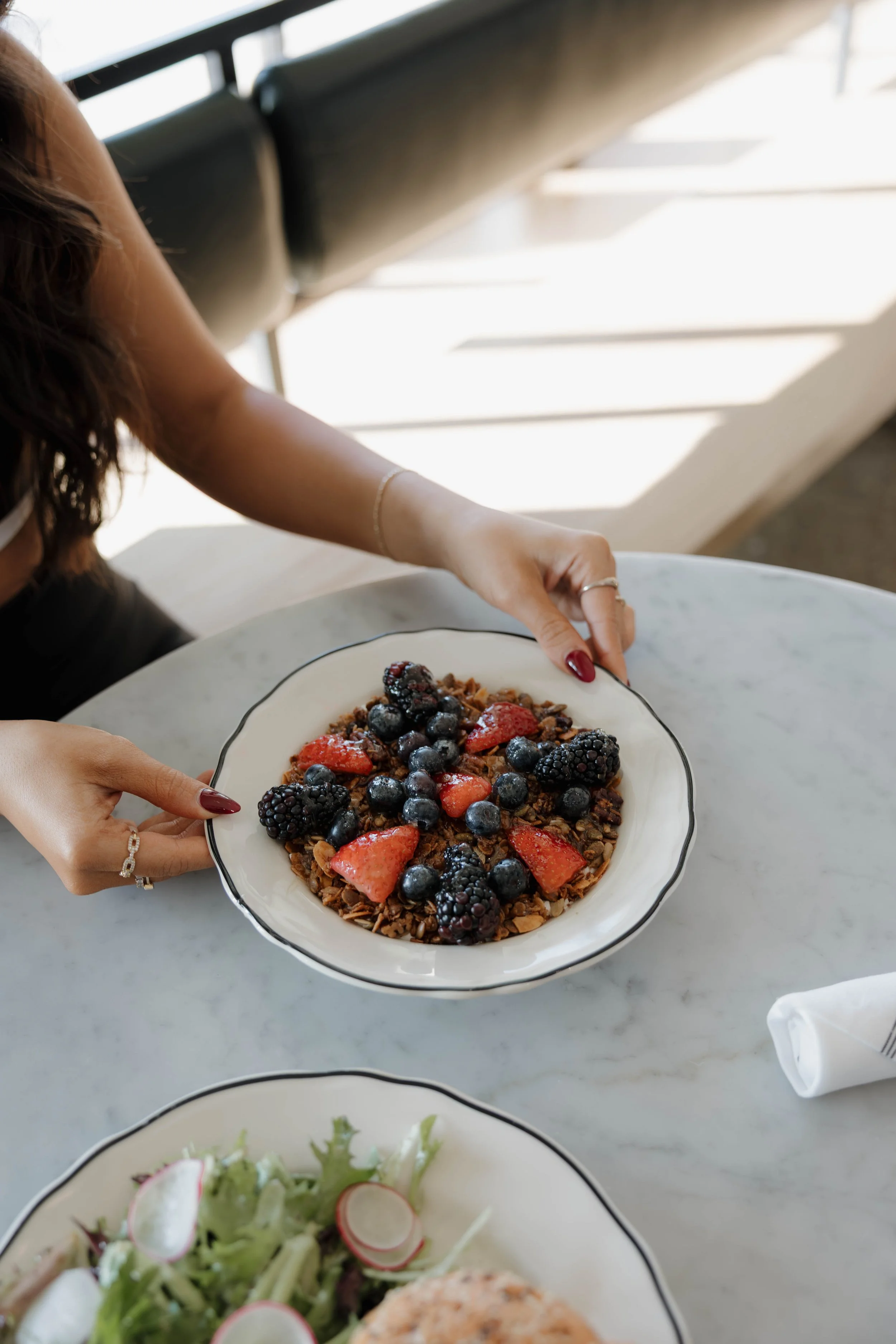 Person holding a bowl of granola topped with mixed berries, including strawberries, blueberries, and blackberries, at a marble table.