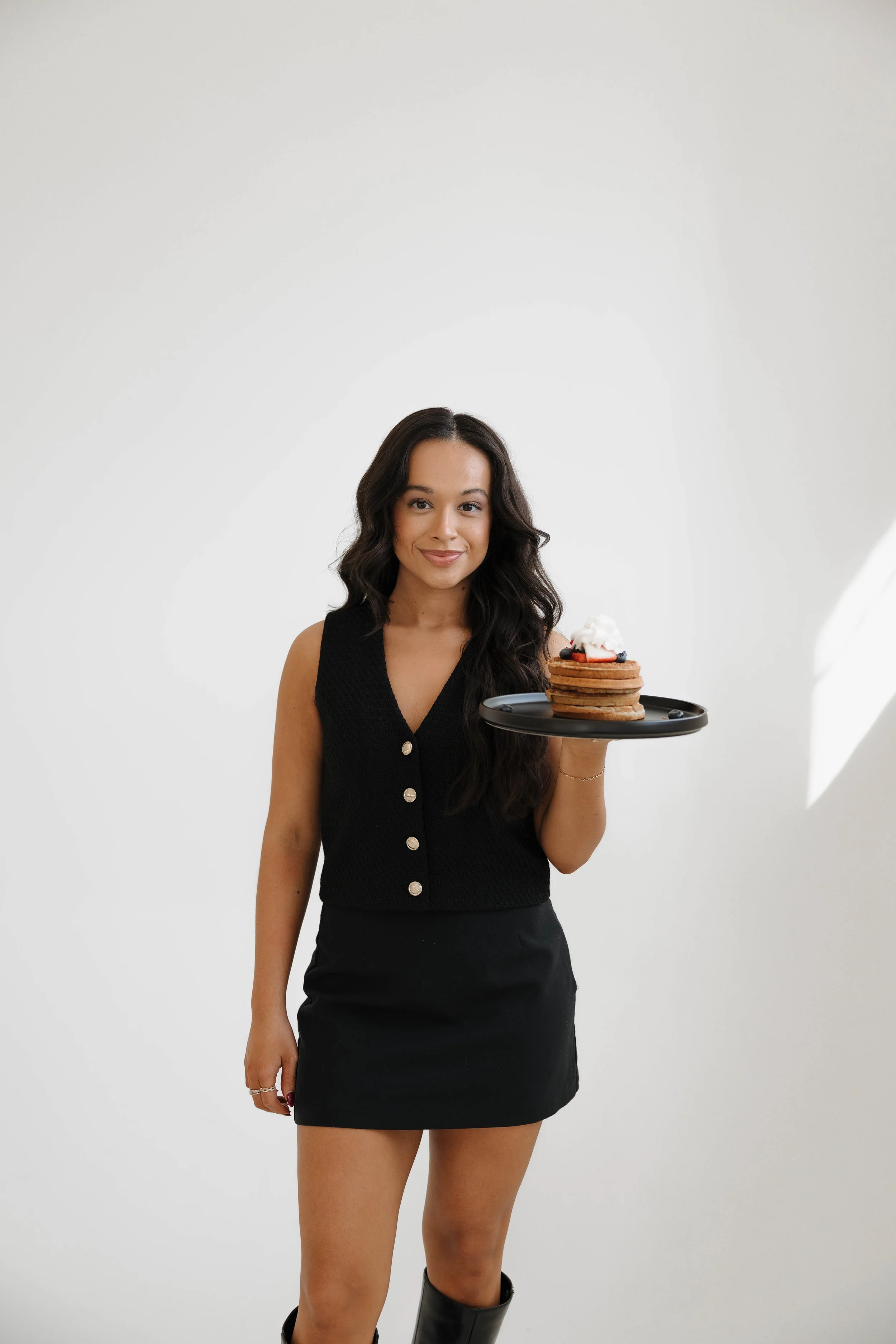 A woman with long dark hair holding a black tray with a stack of pancakes topped with whipped cream and strawberries, standing against a white background.