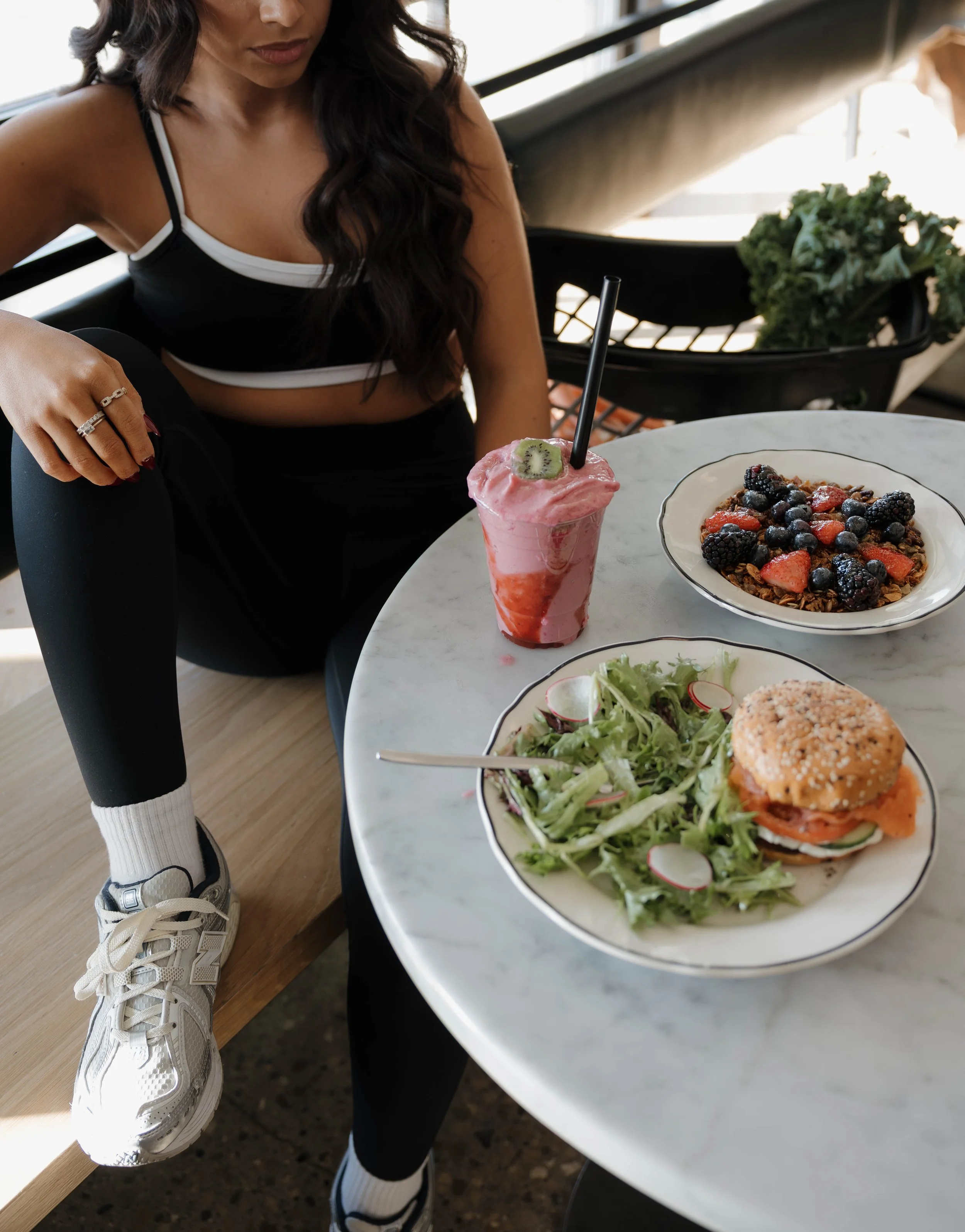 A woman in black workout clothes sitting at a marble table with a pink smoothie, a bowl of granola with berries, a salad with radishes, and a sandwich on a decorative plate.
