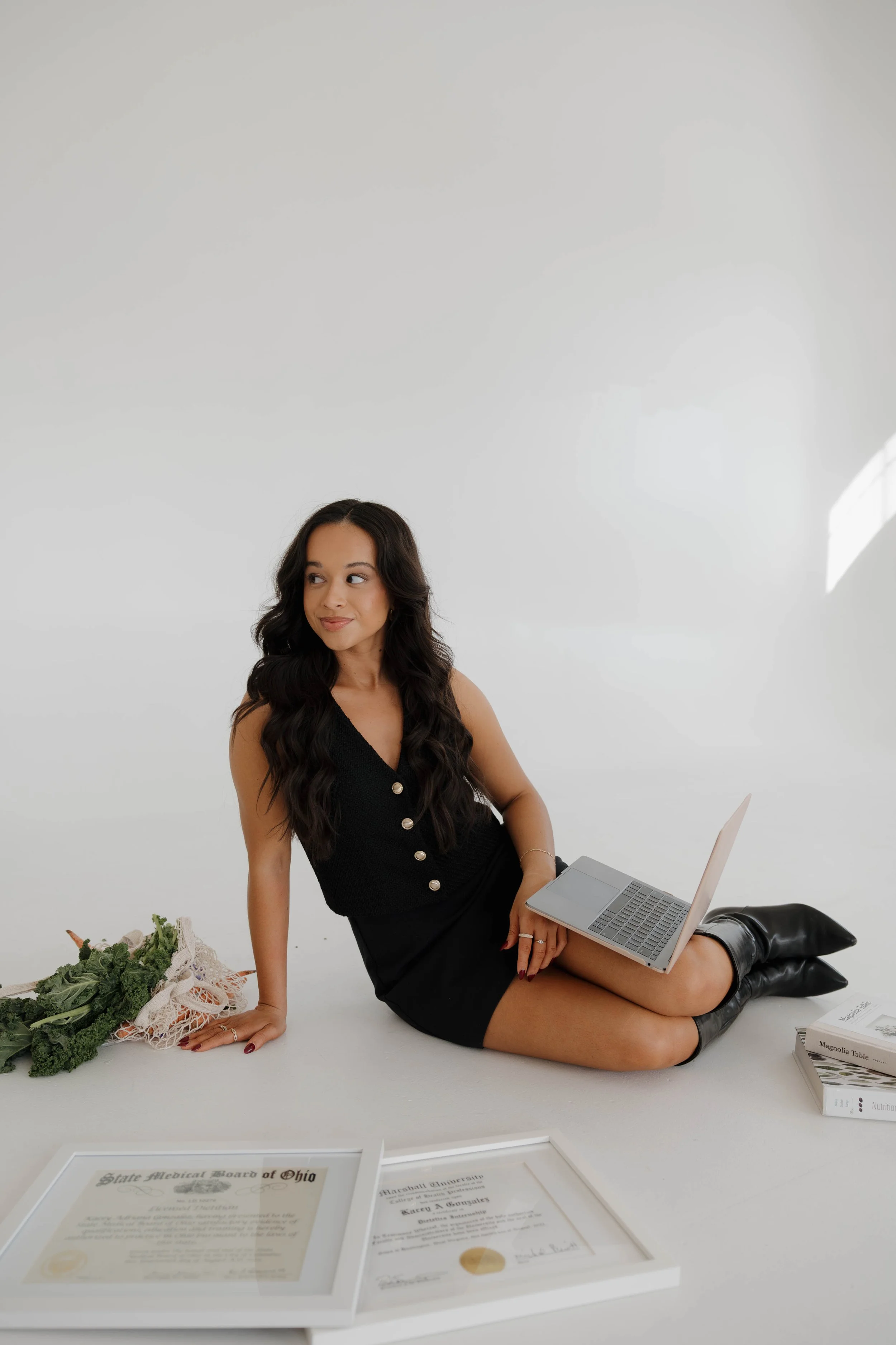 A young woman with dark, wavy hair sitting on the floor with a laptop on her lap, surrounded by documents, books, and vegetables on a white background.
