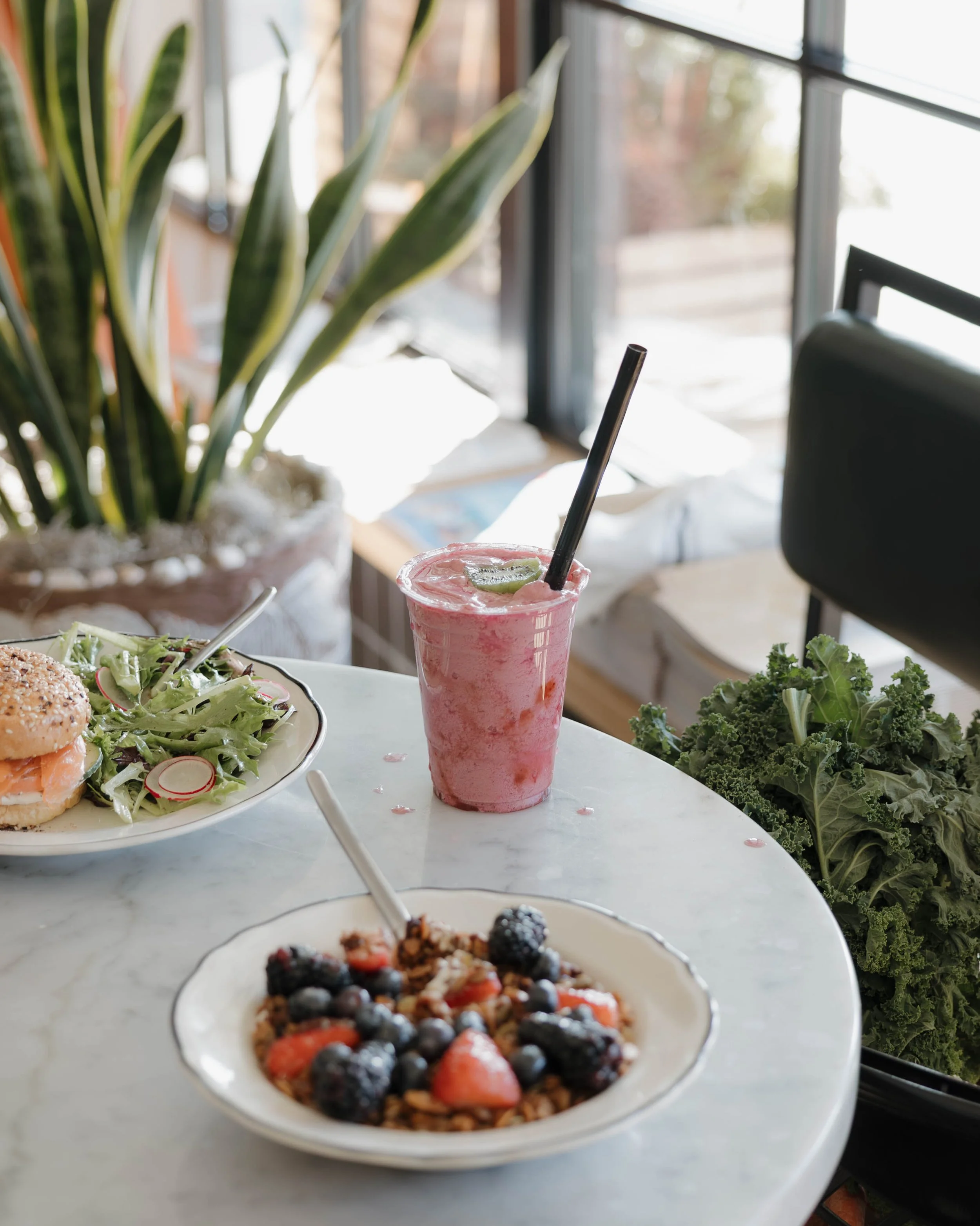 A pink smoothie with a straw and lime slice on top on a marble table, alongside a bowl of mixed berries and granola, a plate of salad, and a plant in the background.