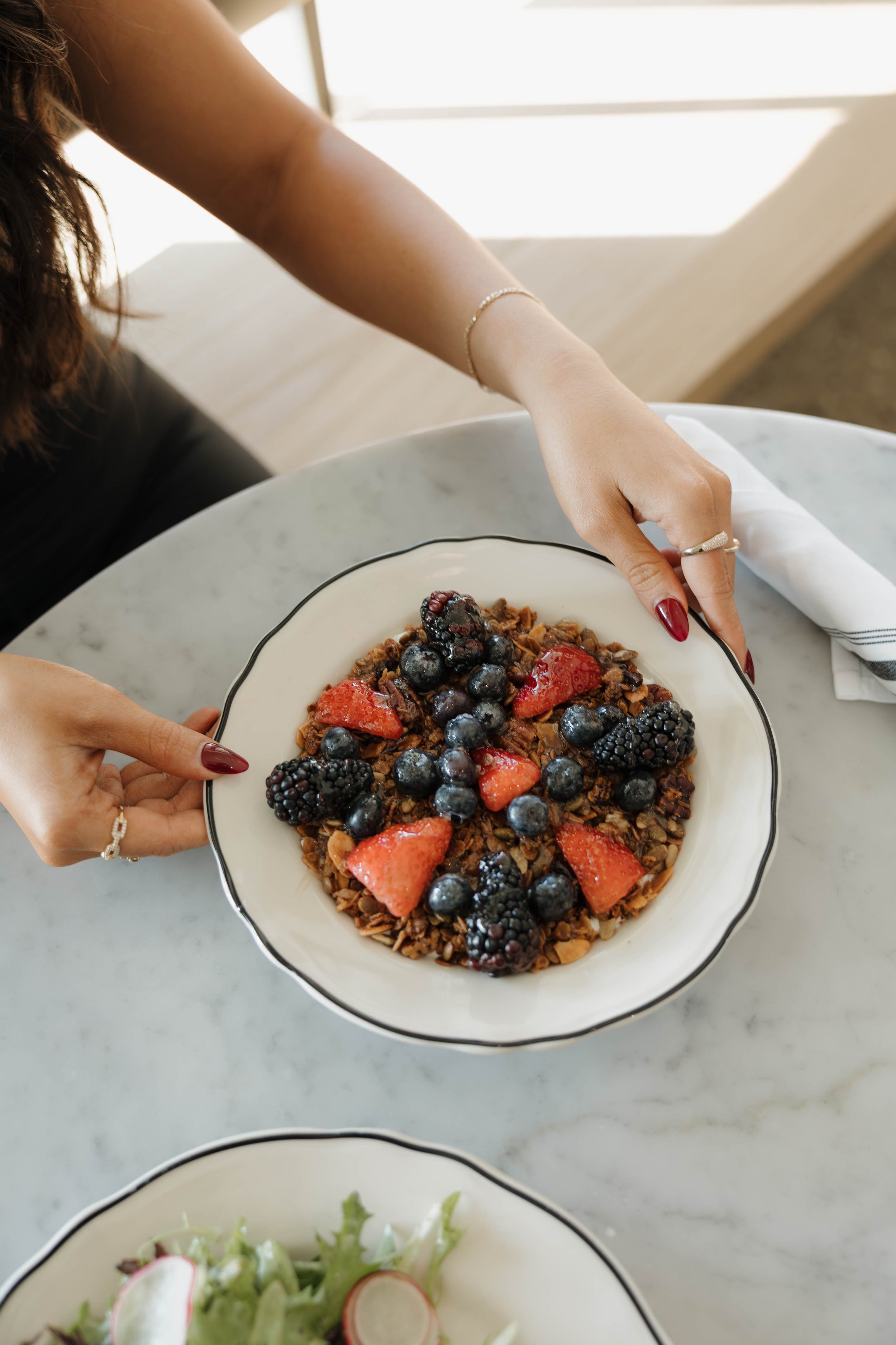 A person is holding a bowl of granola topped with mixed berries, including strawberries, blueberries, and blackberries, on a marble table.