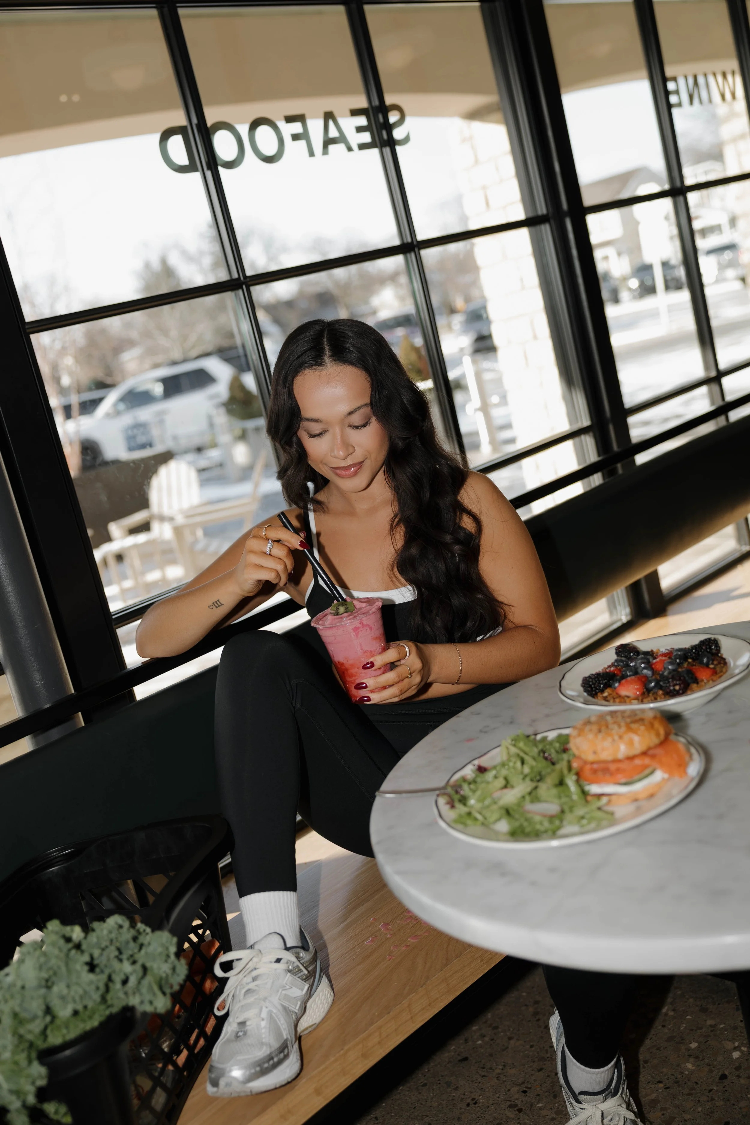 A woman with long black hair sits in a cafe, enjoying a pink blended drink with a straw. She is wearing a black tank top and athletic shoes, with a plate of salad and a sandwich on the marble table beside her. Large windows are behind her, with cars visible outside.