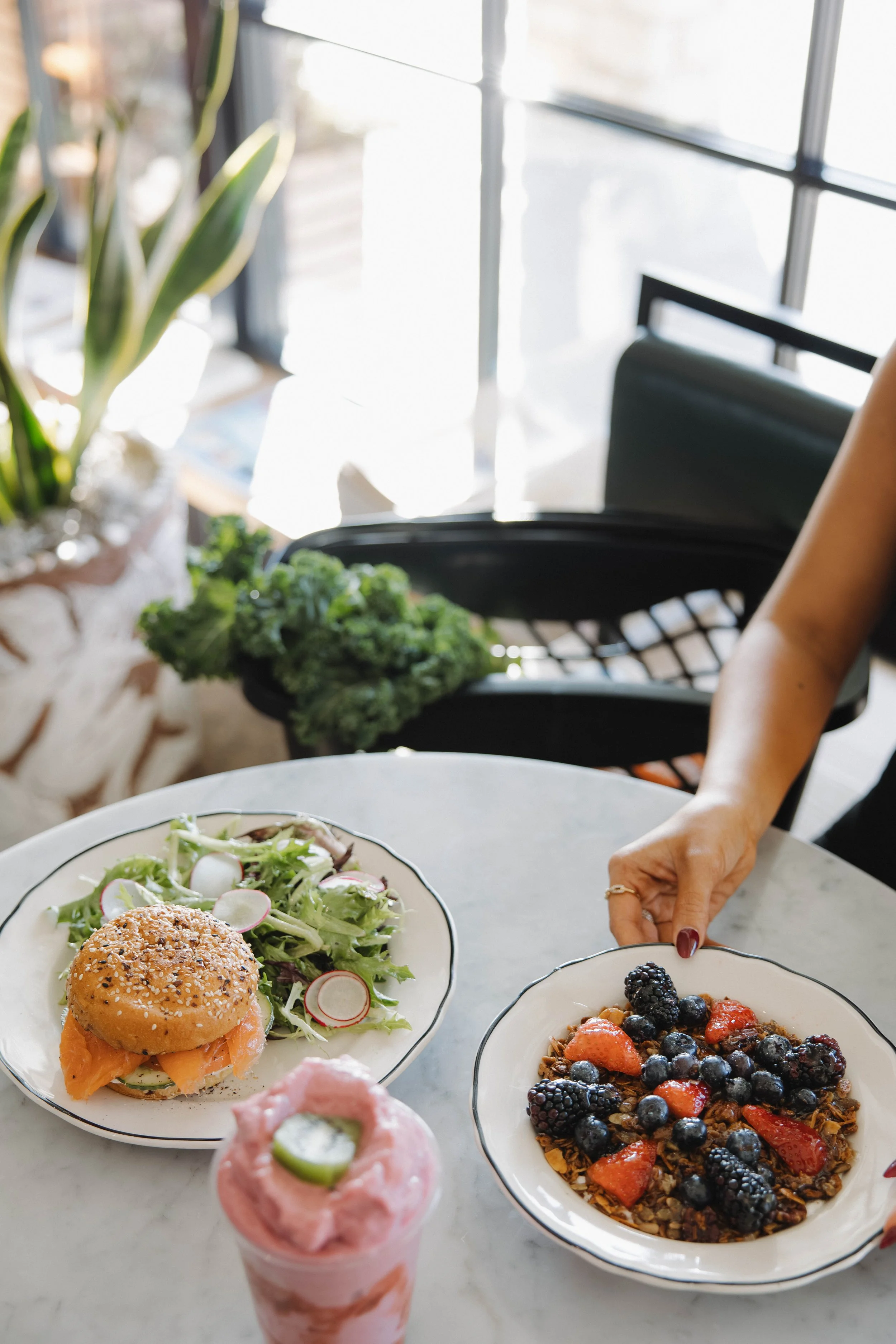 A marble table with a salad, an open-faced smoked salmon sandwich, a bowl of mixed berries and granola, and a pink smoothie garnished with a cucumber slice, with a person reaching for the berry bowl in a bright, cozy cafe.