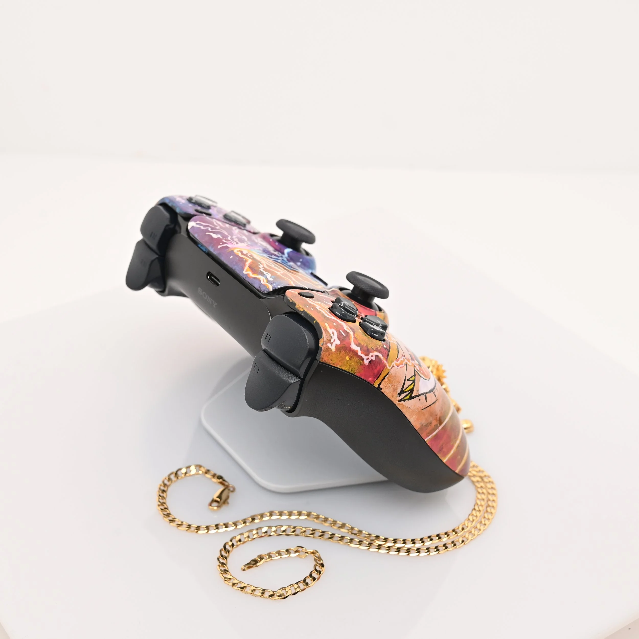 A black gaming controller with colorful, abstract artwork on it, placed on a white stand, with two gold chains in front of it, against a plain white background.