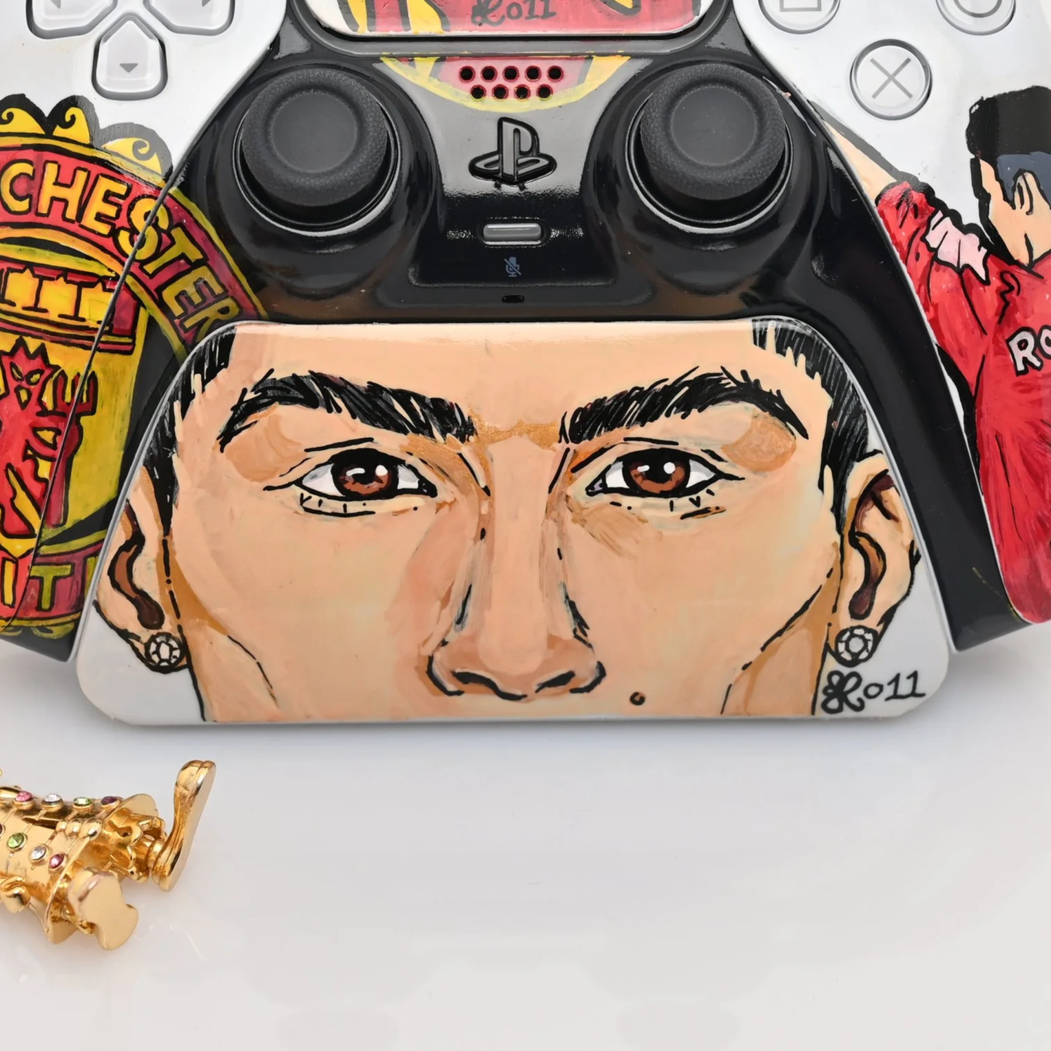 A PlayStation controller with fans art of a woman's face painted on it, showing brown eyes, dark eyebrows, and earrings, surrounded by colorful stickers and a gold ring with gemstones.