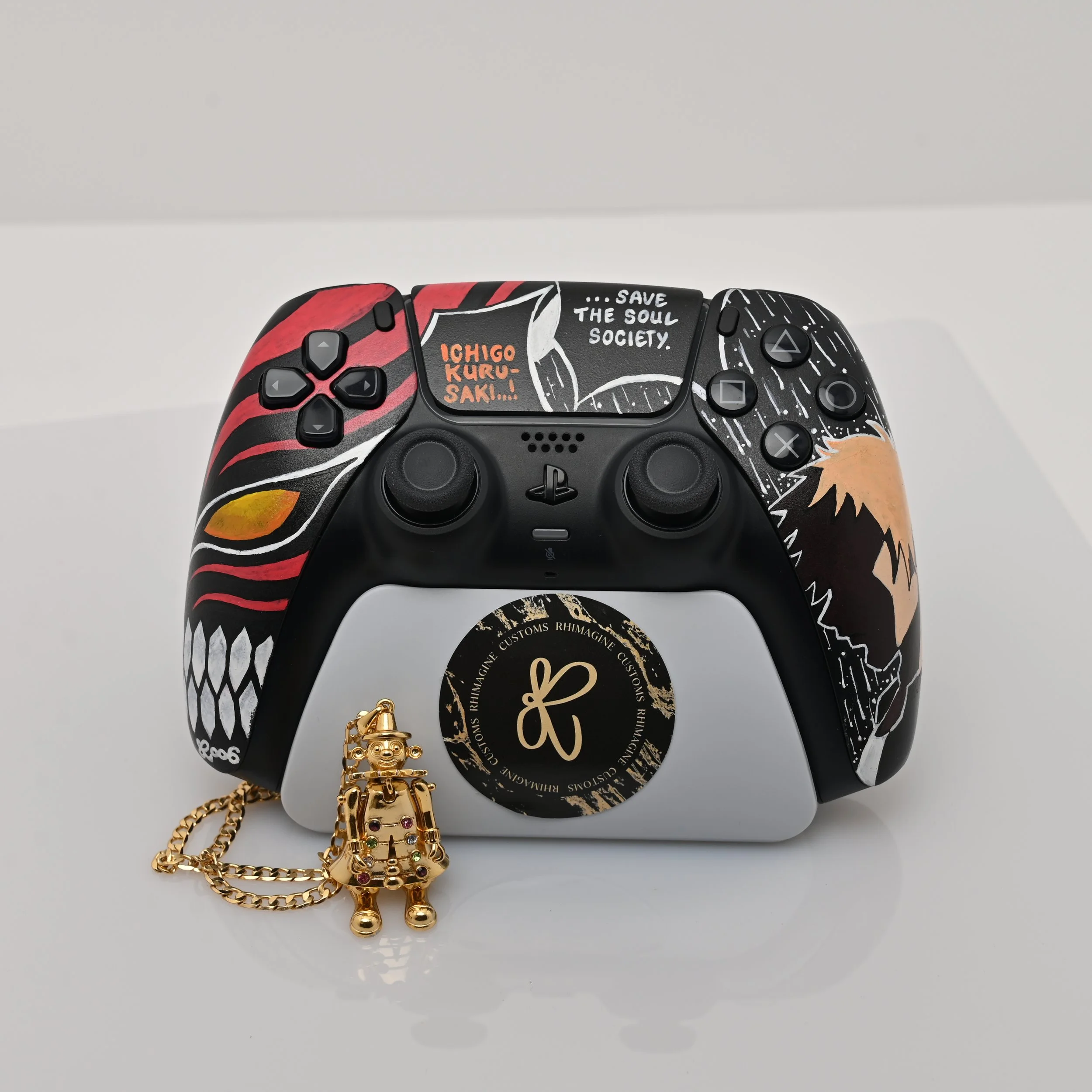 A decorated PlayStation 5 controller with artwork and sayings, alongside a small gold robot figurine keychain with a chain, placed on a white surface against a white background.
