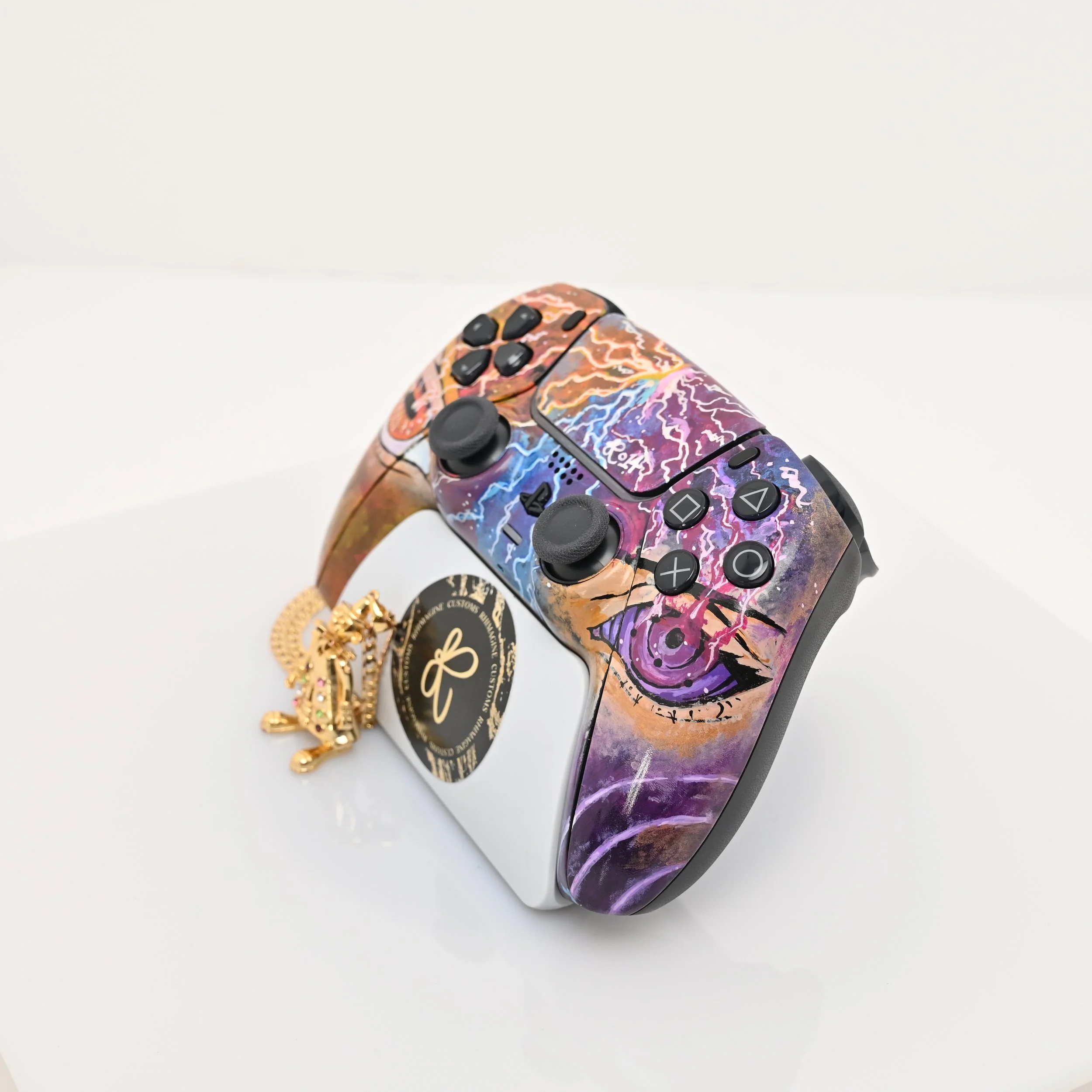 A PlayStation controller with a colorful, abstract lightning design, resting on a white surface. Next to it is a gold chain with a lion pendant and a black circular tag with gold writing.