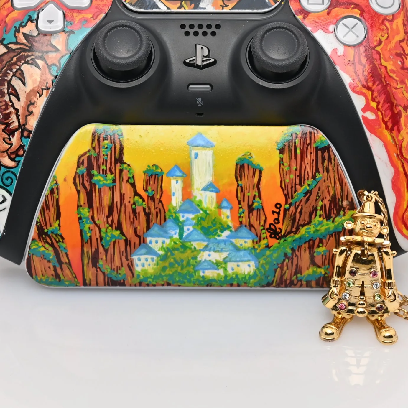A decorated gaming controller with a colorful landscape painting on it, featuring mountains, trees, and a castle, positioned over a white surface with a gold charm figure standing next to it.