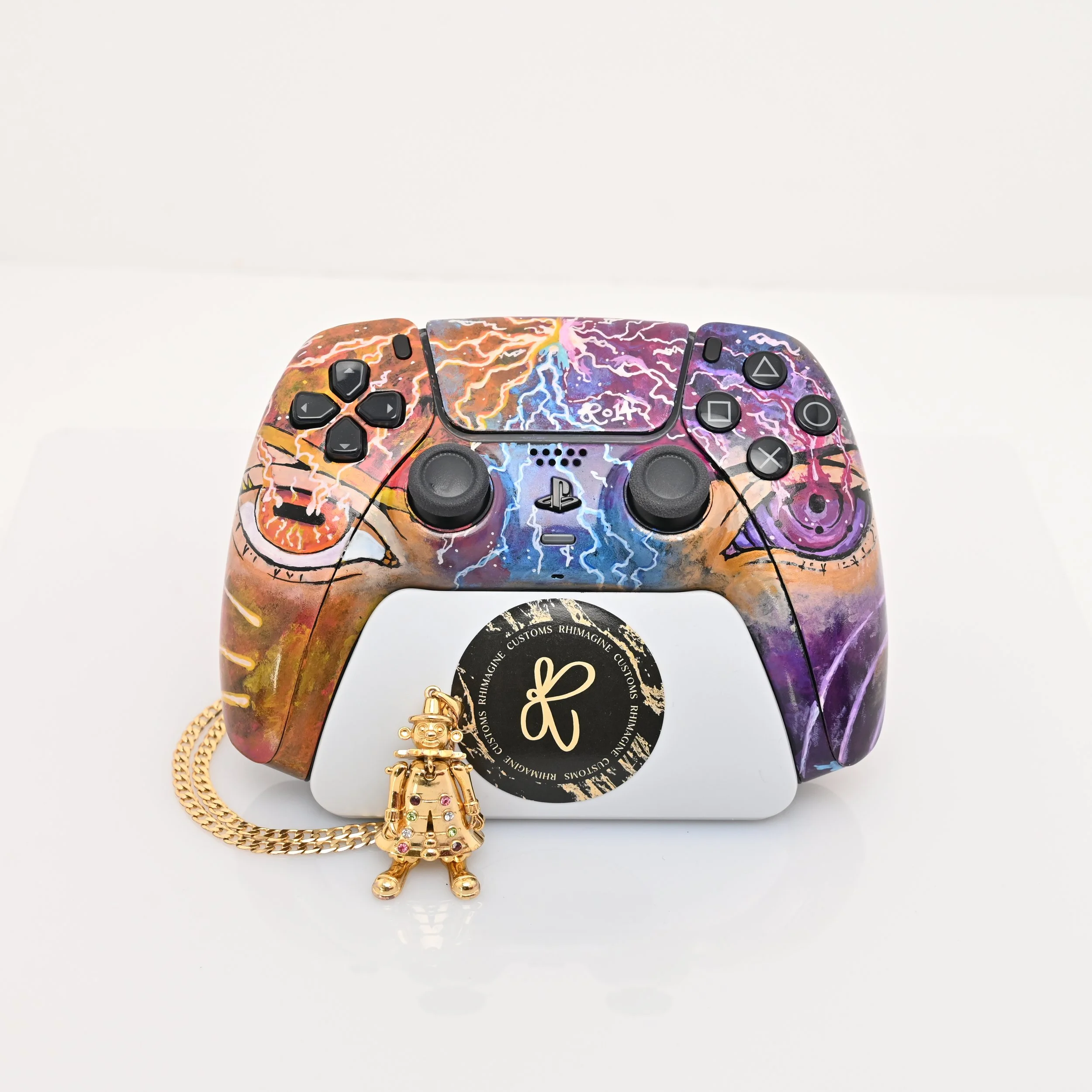 A customized PlayStation controller with a colorful lightning design, placed on a white surface. A gold charm with a bear figure and colorful stones is attached to the controller, and a circular sticker with a black and gold design is on the front.