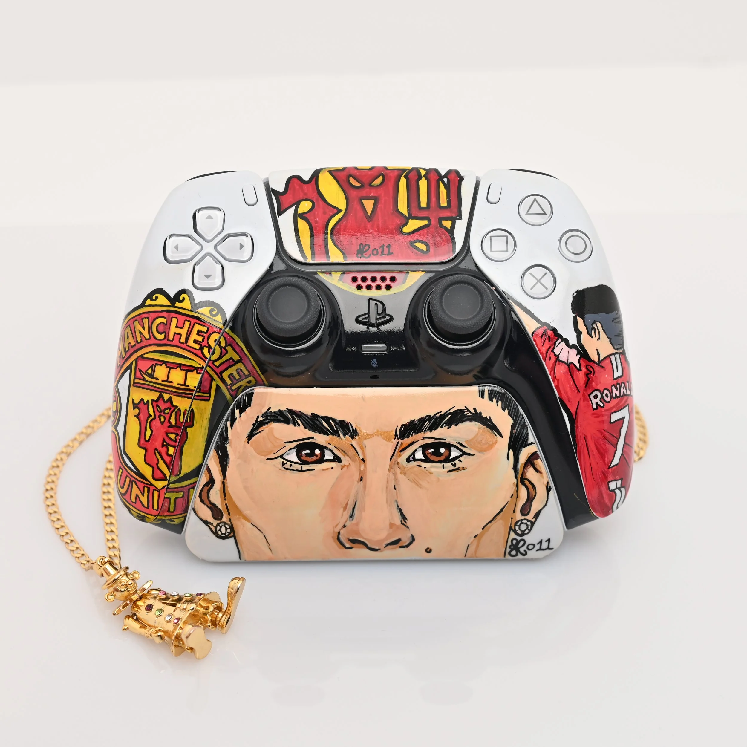Decorated PlayStation controller featuring artwork of a person's face, a Manchester United logo, a depiction of Ronaldinho in a red jersey, and a collage of red and yellow elements, with a gold chain and a small king figurine necklace.