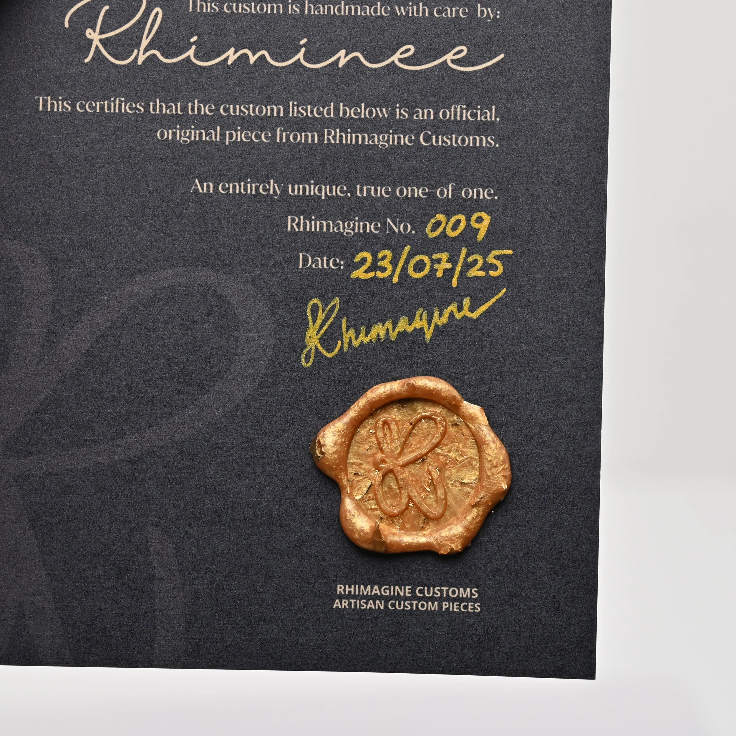 A certificate from Rhimagine Customs authenticating a handmade, original piece with a signature, along with a wax seal featuring a stylized heart. The certificate includes details such as Rhiaminee, Rhiaminee Customs, Rhiaminee No. 009, date 23/07/25, and a signature in gold ink.