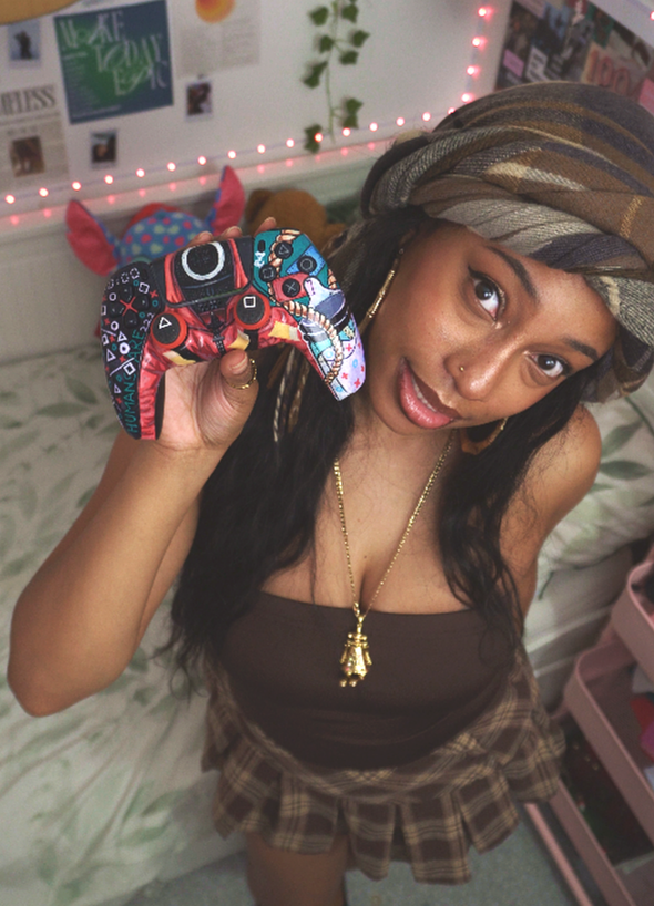 A woman with long dark hair, wearing a plaid skirt, a brown top, a gold chain and a gold pendant, is holding a colorful video game controller. She is smiling and tilting her head to the side, with a headscarf on her head and a decorated room background.