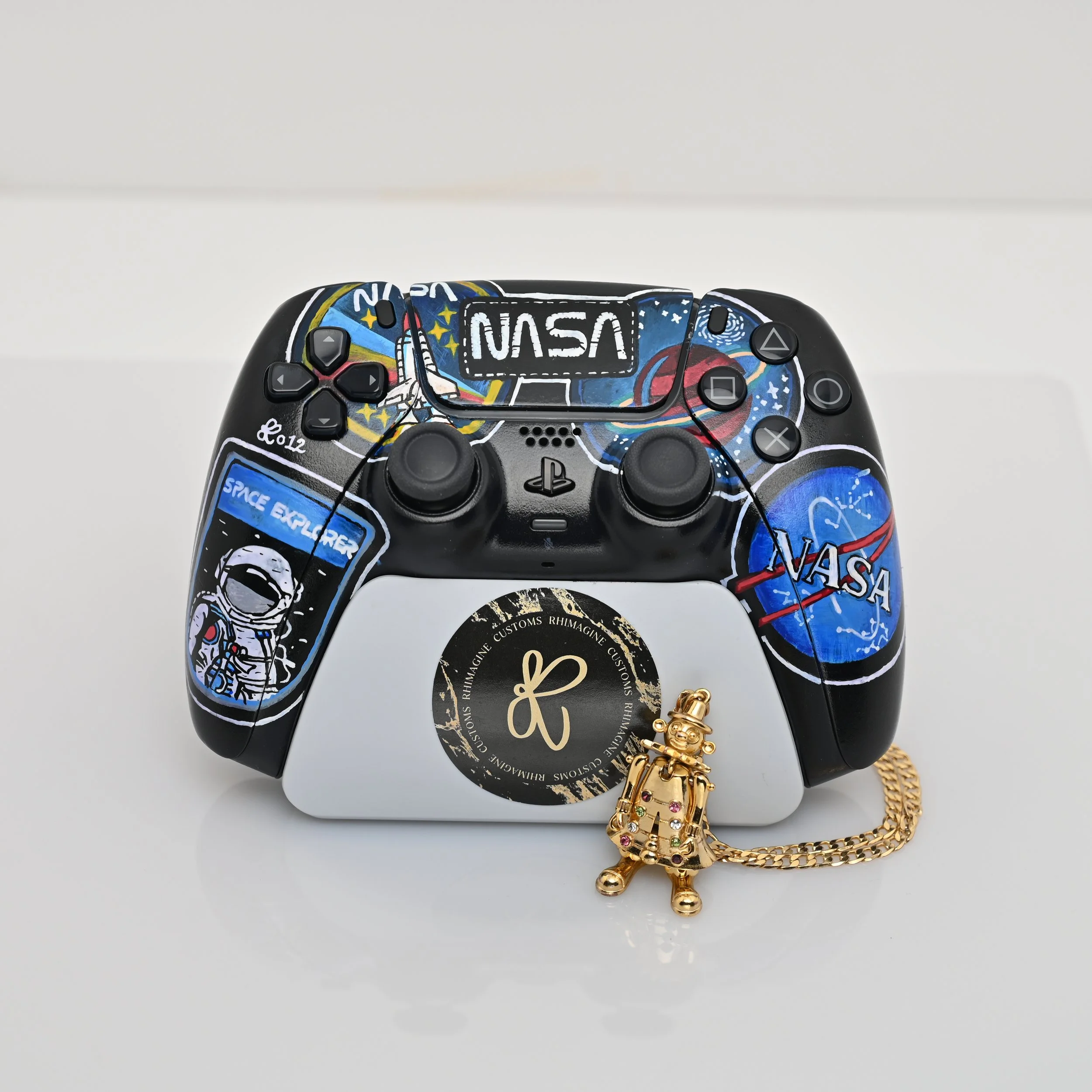 A custom PlayStation controller decorated with NASA-themed illustrations, featuring the NASA logo, astronaut, and space-themed elements. In front of the controller is a gold chain with a small astronaut figurine pendant. The controller is placed on a