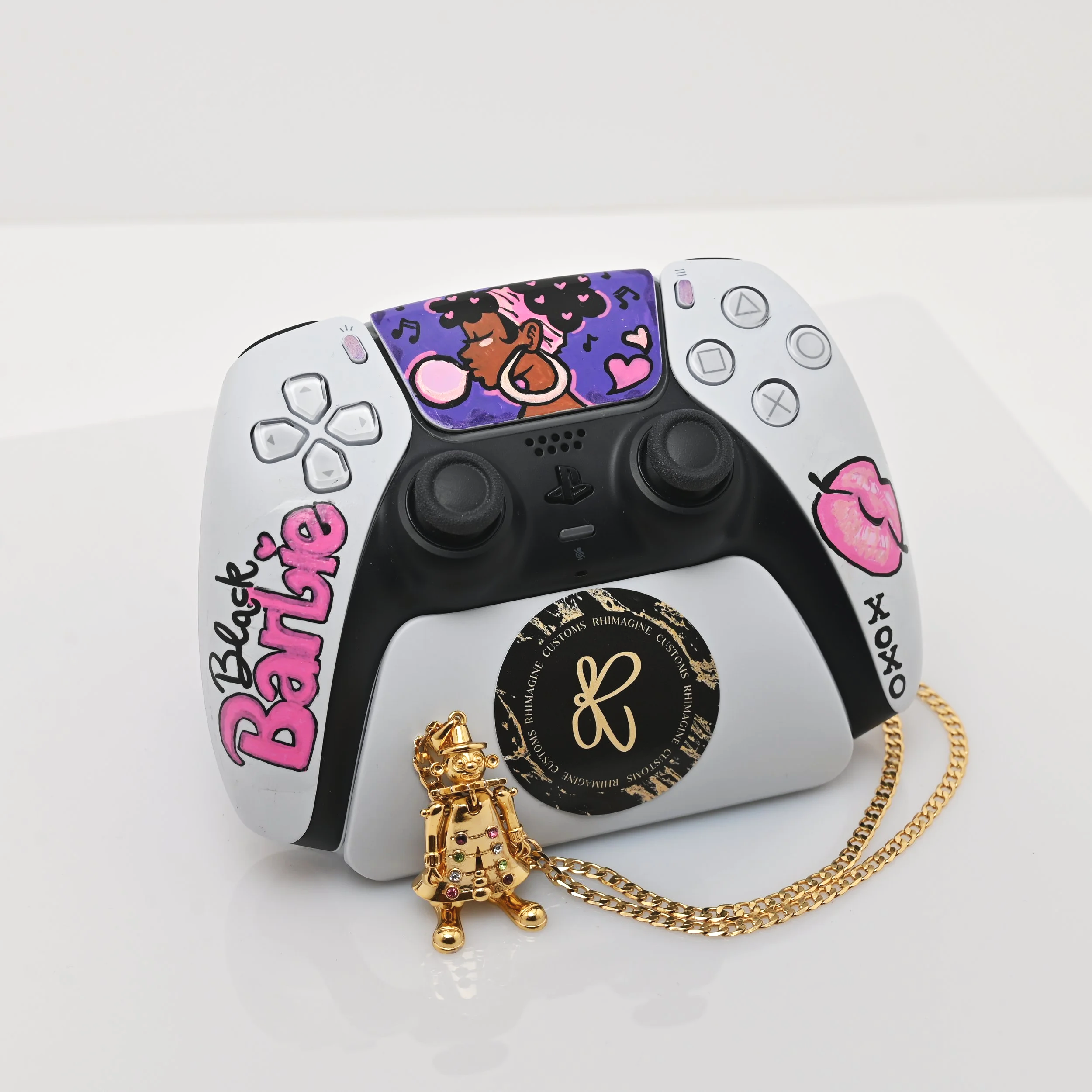 A decorated PlayStation 5 controller with stickers, including a cartoon girl, pink lips, and pink text reading 'Back Barbie', placed on a white surface with gold jewelry in front.