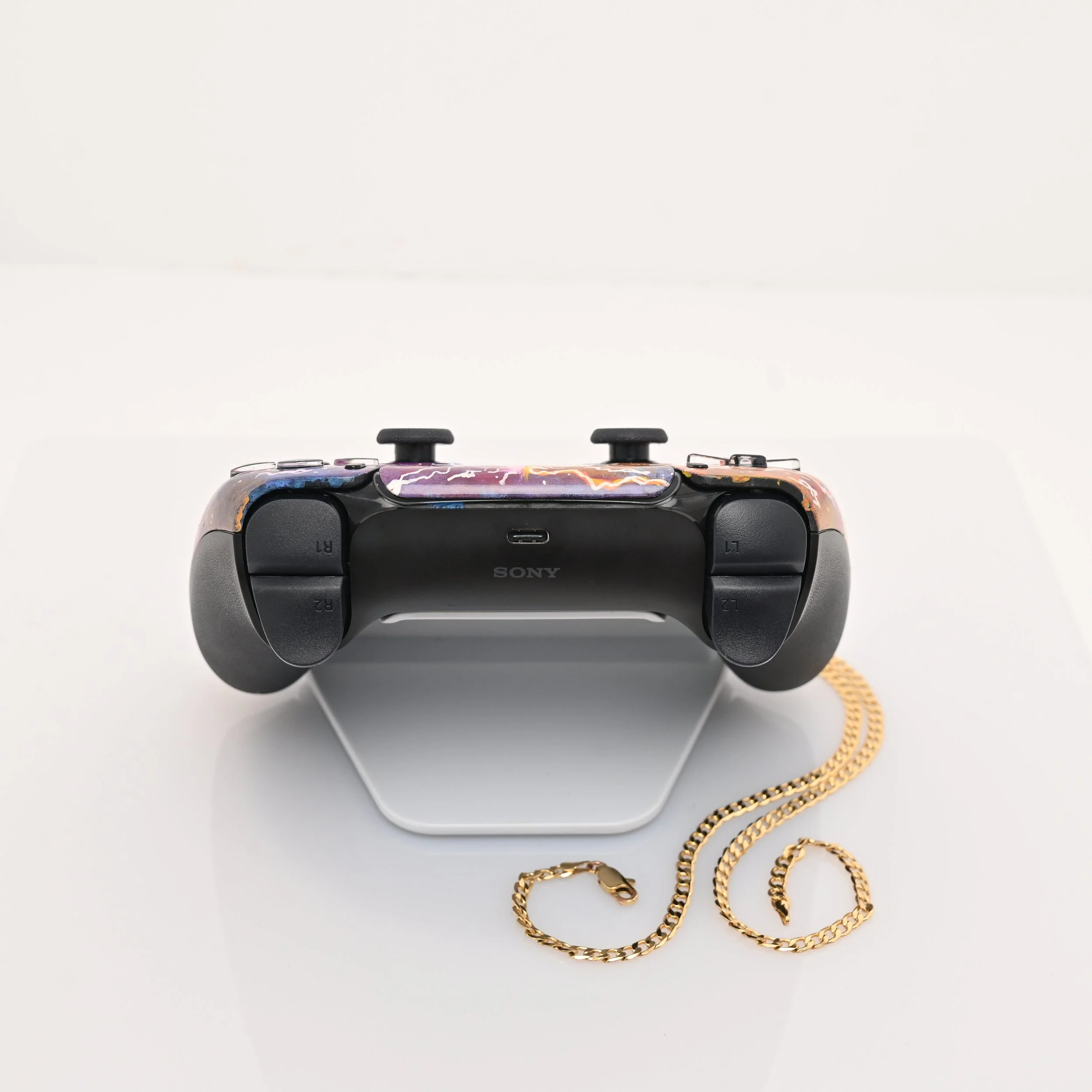 A Sony gaming controller with a colorful, lightning-themed design on the top, resting on a white stand, with a gold chain placed in front of it on a white surface.