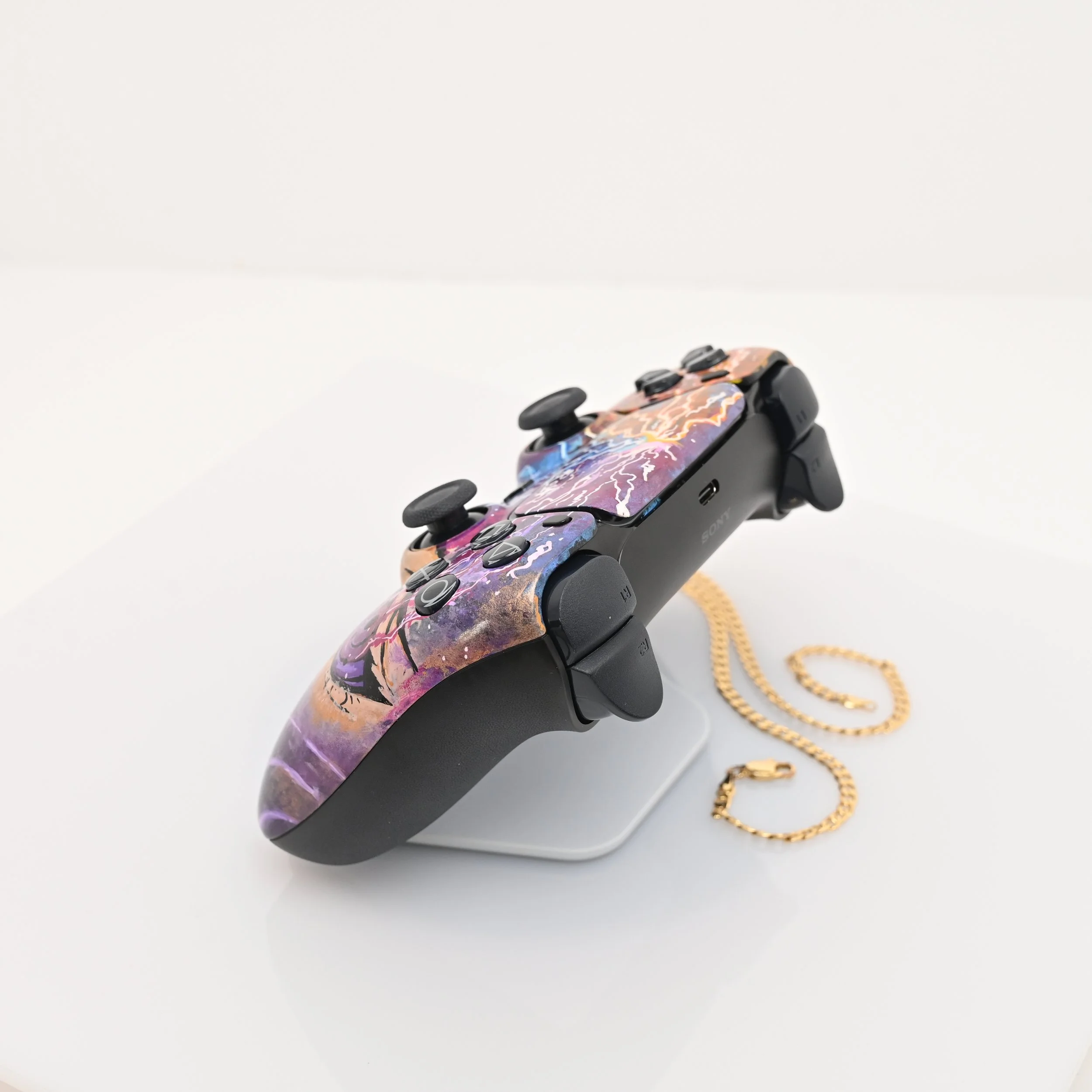 A black PlayStation controller decorated with a colorful and abstract art print, placed on a stand with a gold chain accessory nearby.