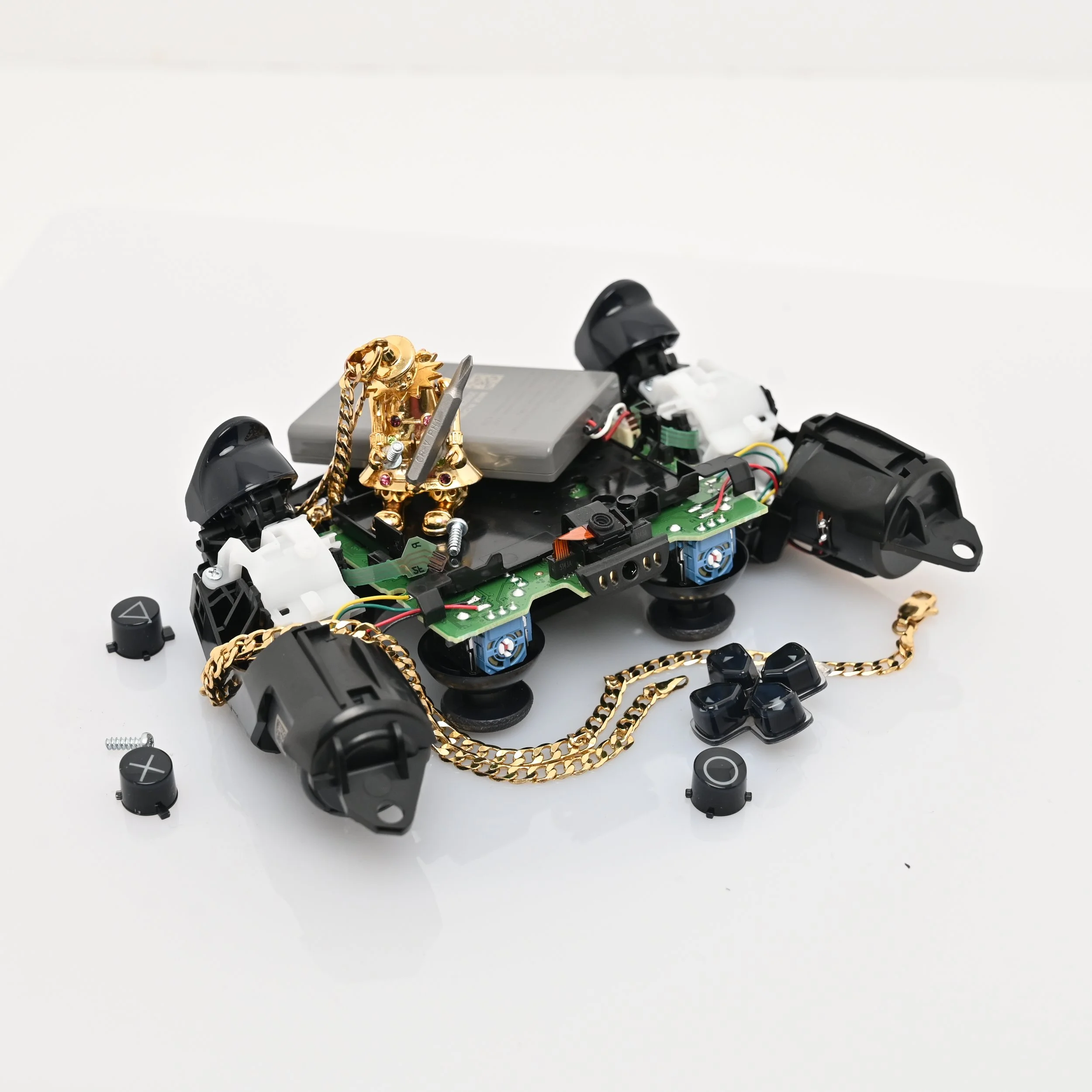 Disassembled gaming controller with circuit board, joysticks, buttons, and gold chains on a white surface.