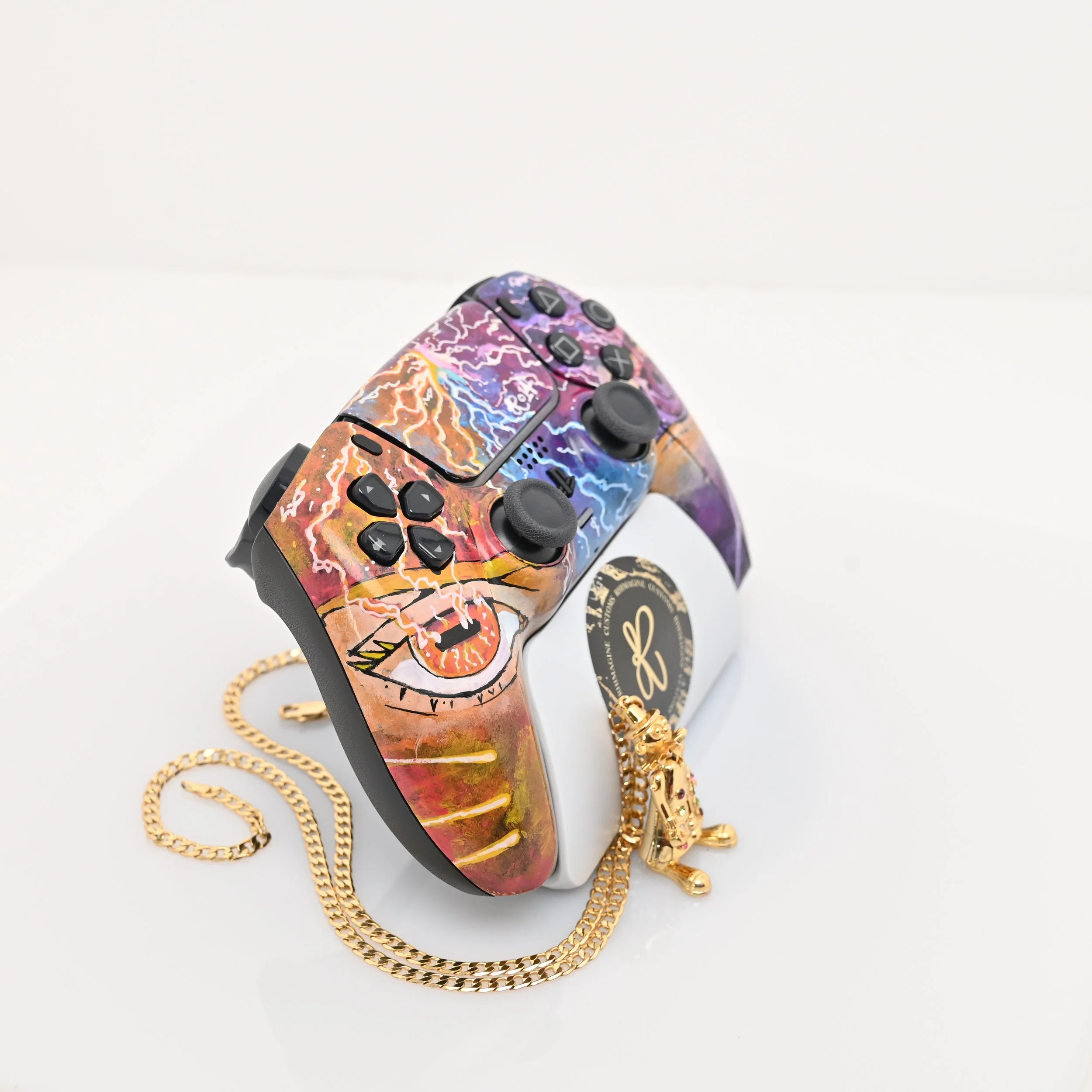 A video game controller with a colorful lightning storm design, placed on a white surface with a gold chain necklace and a small decorative lion figurine nearby.