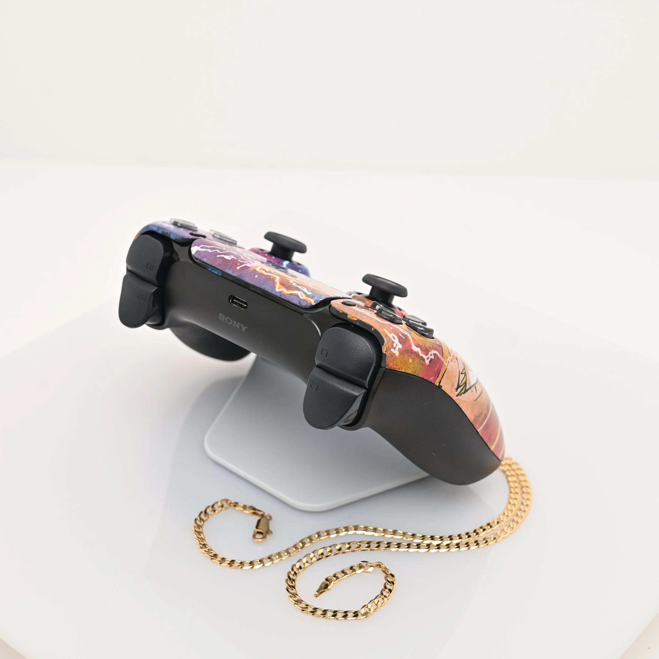 A PlayStation controller with a colorful, abstract design on top, resting on a white display stand with a gold chain in front.
