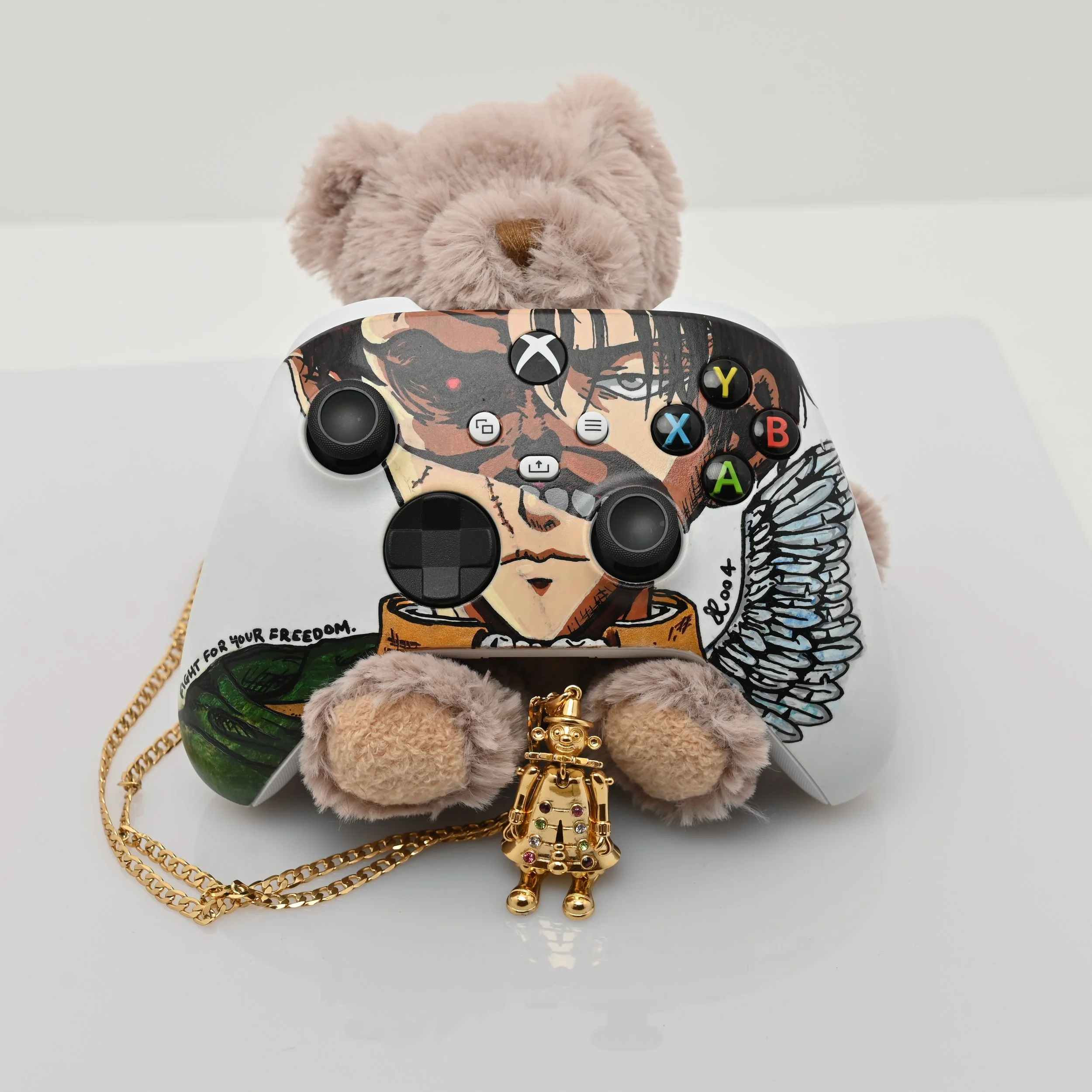 A custom Xbox controller with levi ackerman from attack on titan with a beast titan, surrounded a wing, placed on a white surface. Behind the controller is a brown teddy bear, and below is a small gold clown charm attached to a gold chain. 