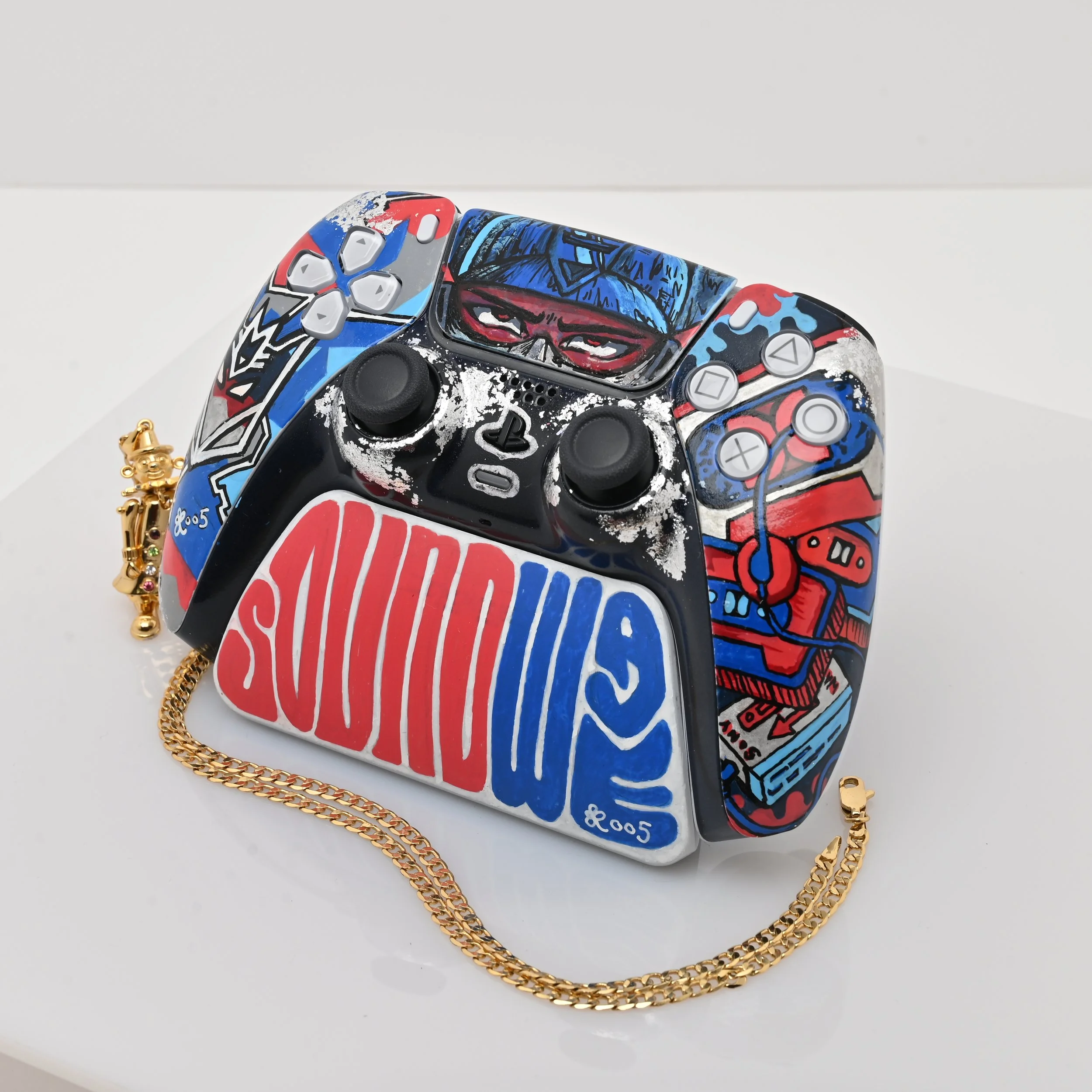 A colorful handbag designed to look like a vintage PlayStation controller with graffiti-style art, including a face with sunglasses and a cap, and a gold chain strap resting on a white surface.