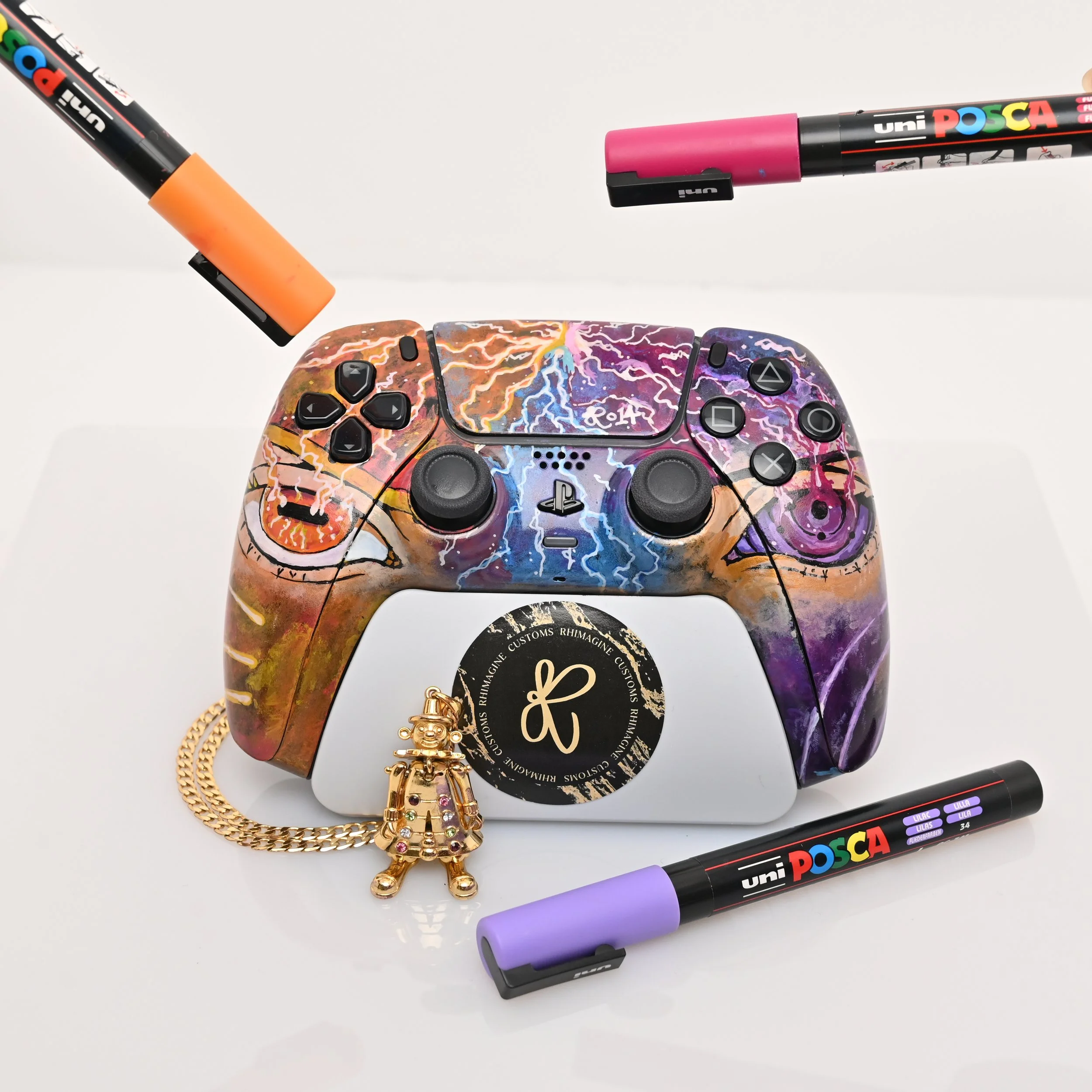 A decorated PlayStation 5 controller with colorful lightning designs, surrounded by four Uni POSCA markers, a small gold chain, and a gold figurine keychain with rhinestones, all on a white surface.
