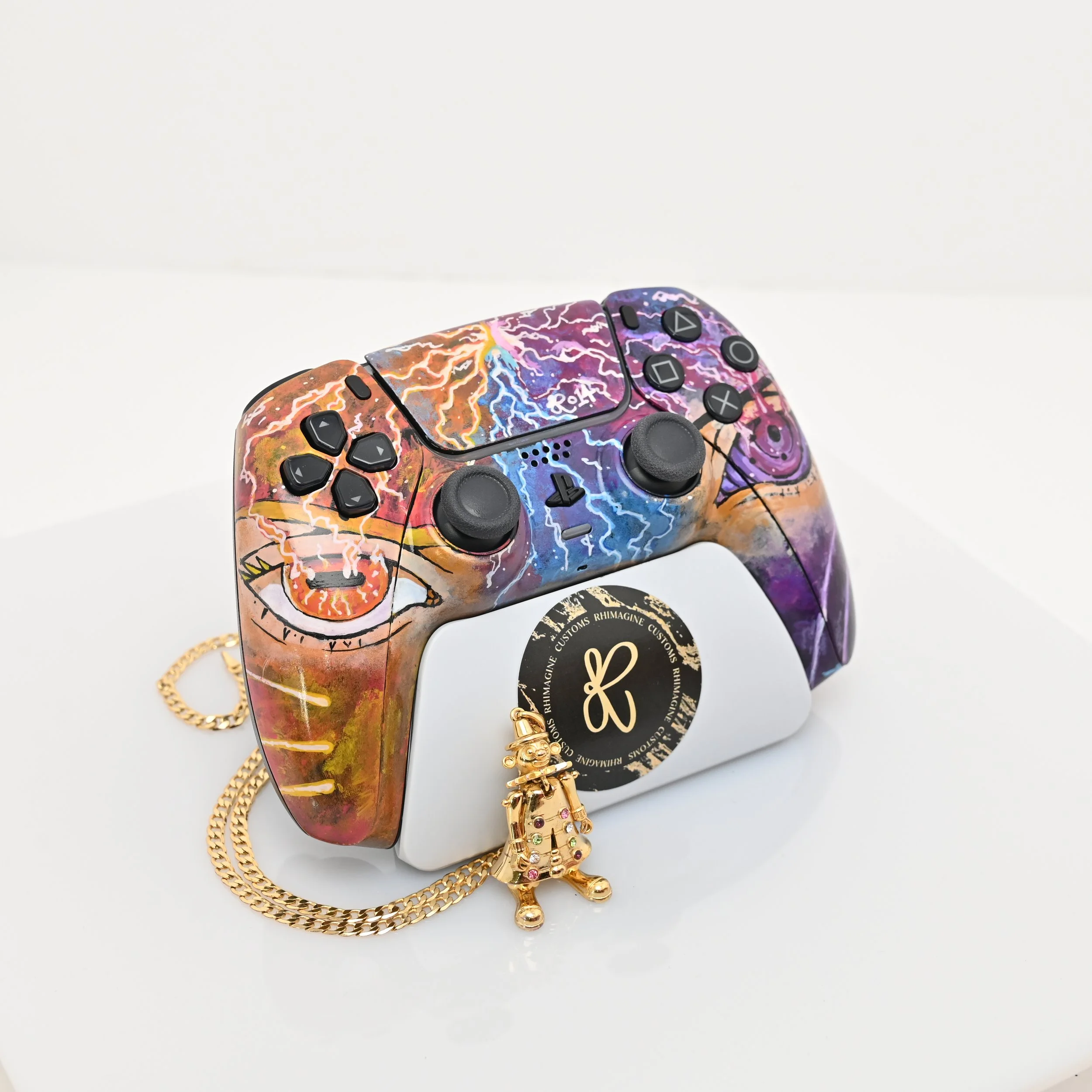 Colorful custom-painted PlayStation controller resting on a white box, adorned with a gold chain and a small gold charm, against a plain white background.