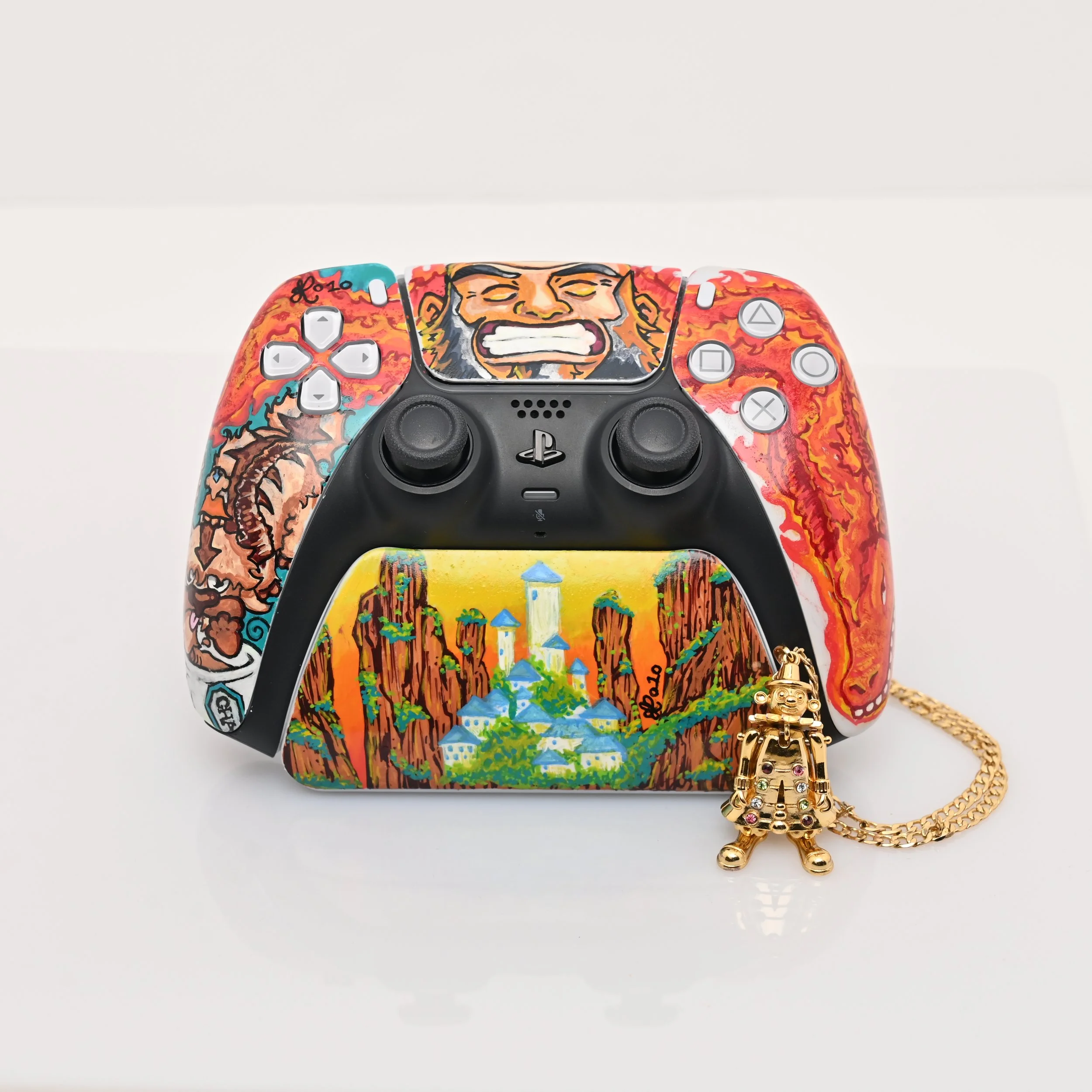 A custom painted PlayStation 5 controller with a colorful mountain landscape and a fierce face on the top. Beside it is a small gold figurine keychain with a chain, featuring a robot-like character with gemstones embedded.