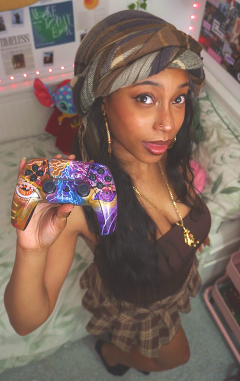 A young woman with dark curly hair wearing a brown headscarf, gold jewelry, and a brown outfit, holding a colorful gaming controller, standing in a bedroom with posters and string lights in the background.