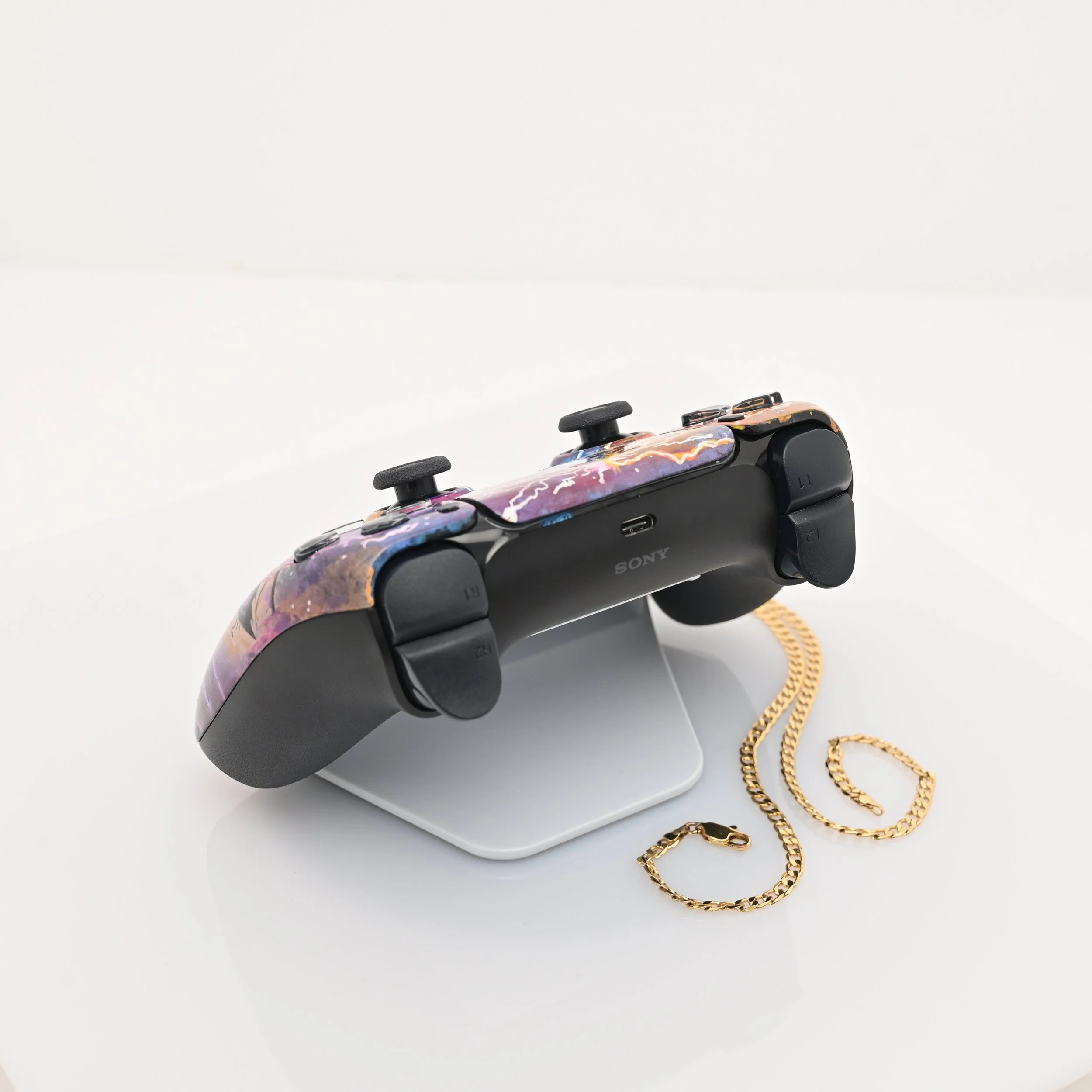 A Sony gaming controller with a colorful, galaxy-like design is displayed on a stand, accompanied by a gold chain placed on a white surface.