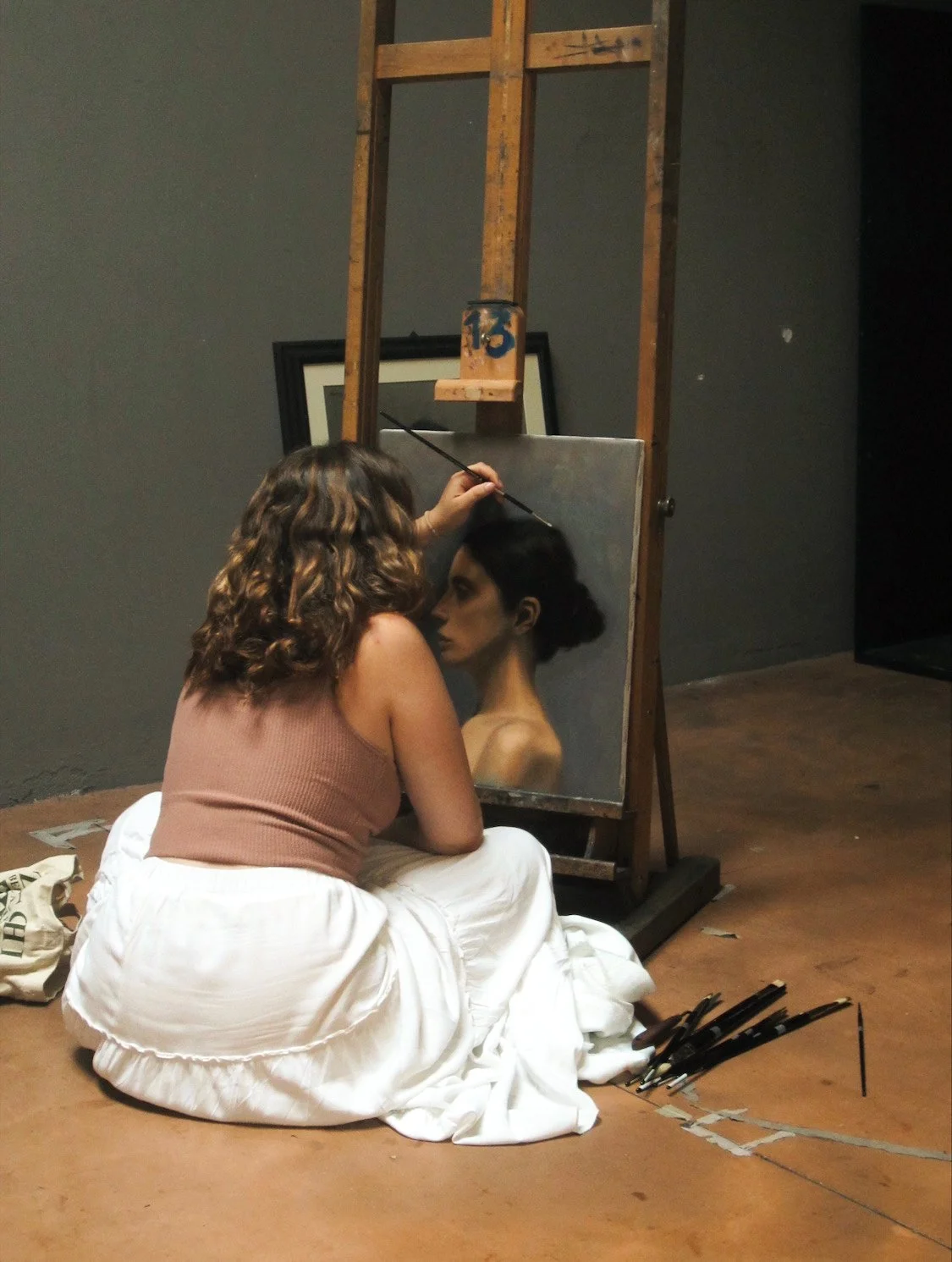 Artist isabel couceiro sits on the floor in front of an easel. she is painting a portrait of a woman's profile in a classical style.