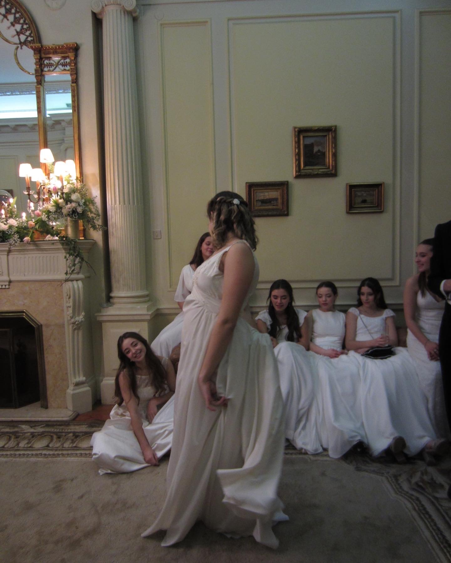 an artistic photograph of young debutante girls in flowing white gowns. One girl in the middle is flaunting her dress and the 5 girls behind her sit on a couch and admire her with smiles.