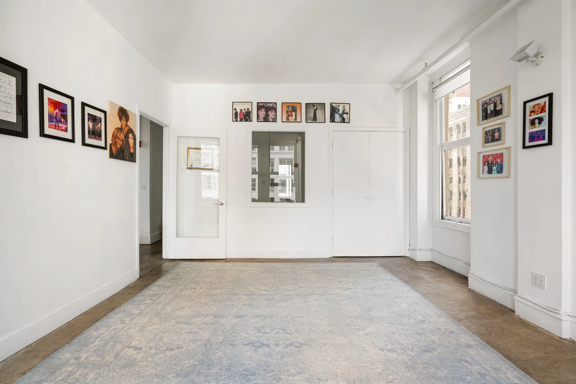 A bright art gallery with white walls displaying framed photographs and artwork, a large window on the right, and a light-colored rug on the wooden floor.