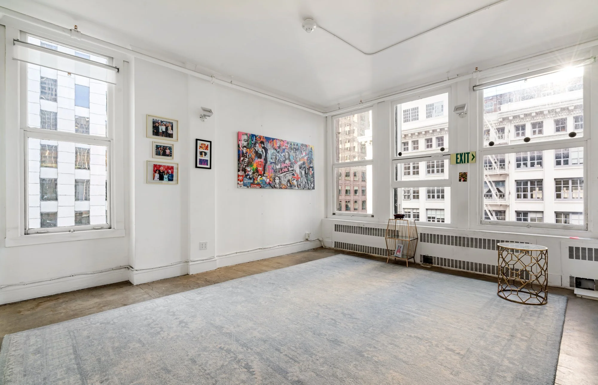 Empty art gallery room with white walls, large windows with cityscape view, artwork on walls, and minimal furniture.