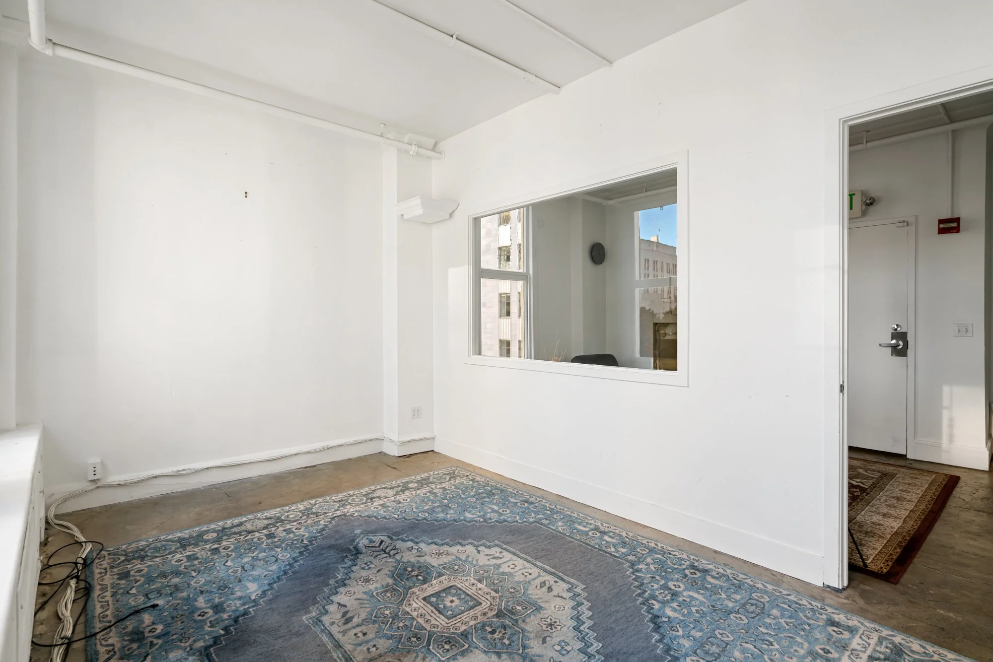 Empty room with white walls, a patterned blue and gray rug, a window with views of neighboring buildings, and a doorway leading to another space.