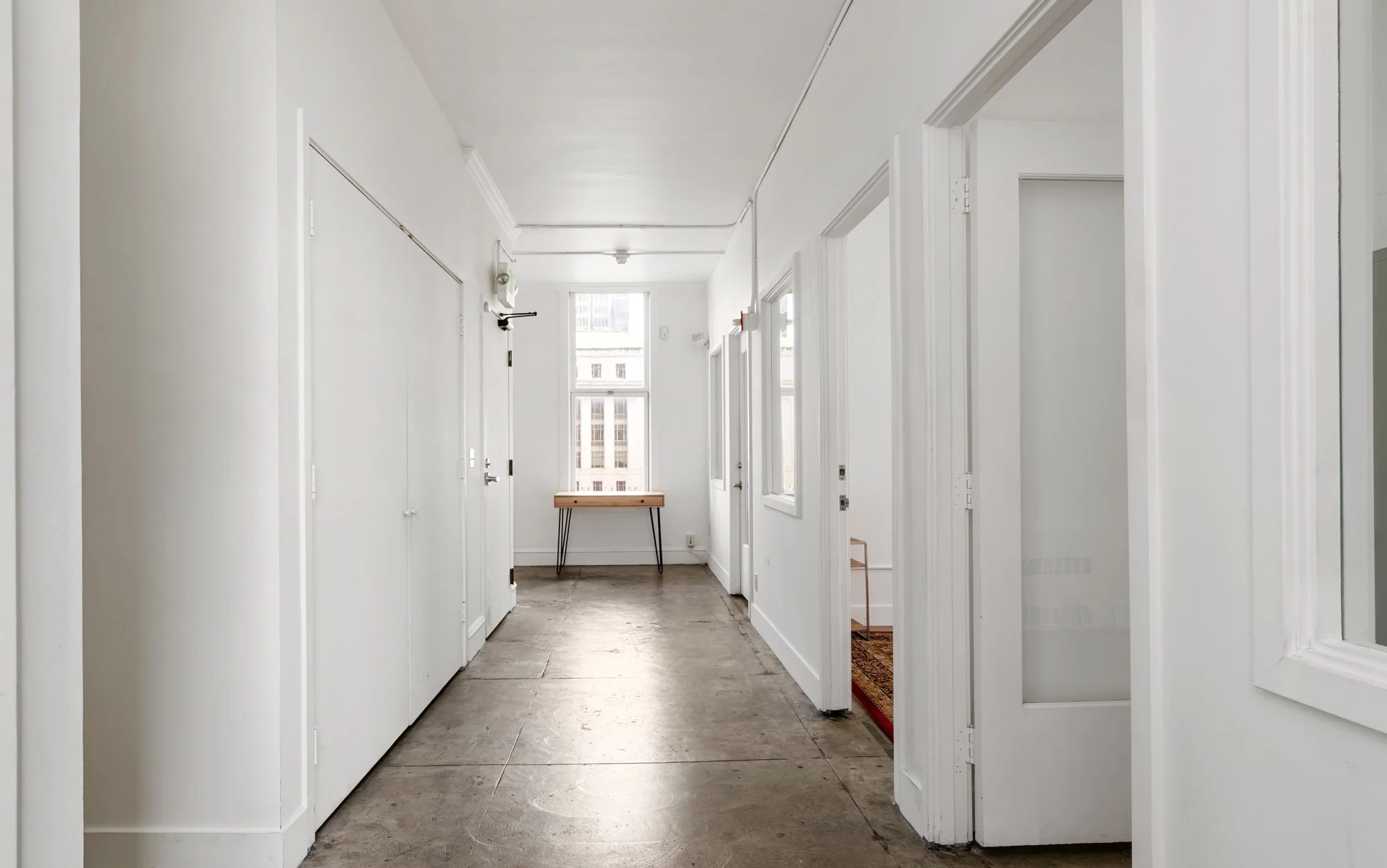 Empty white hallway with concrete floor, several doorways, a window at the end, and a wooden table beneath the window.