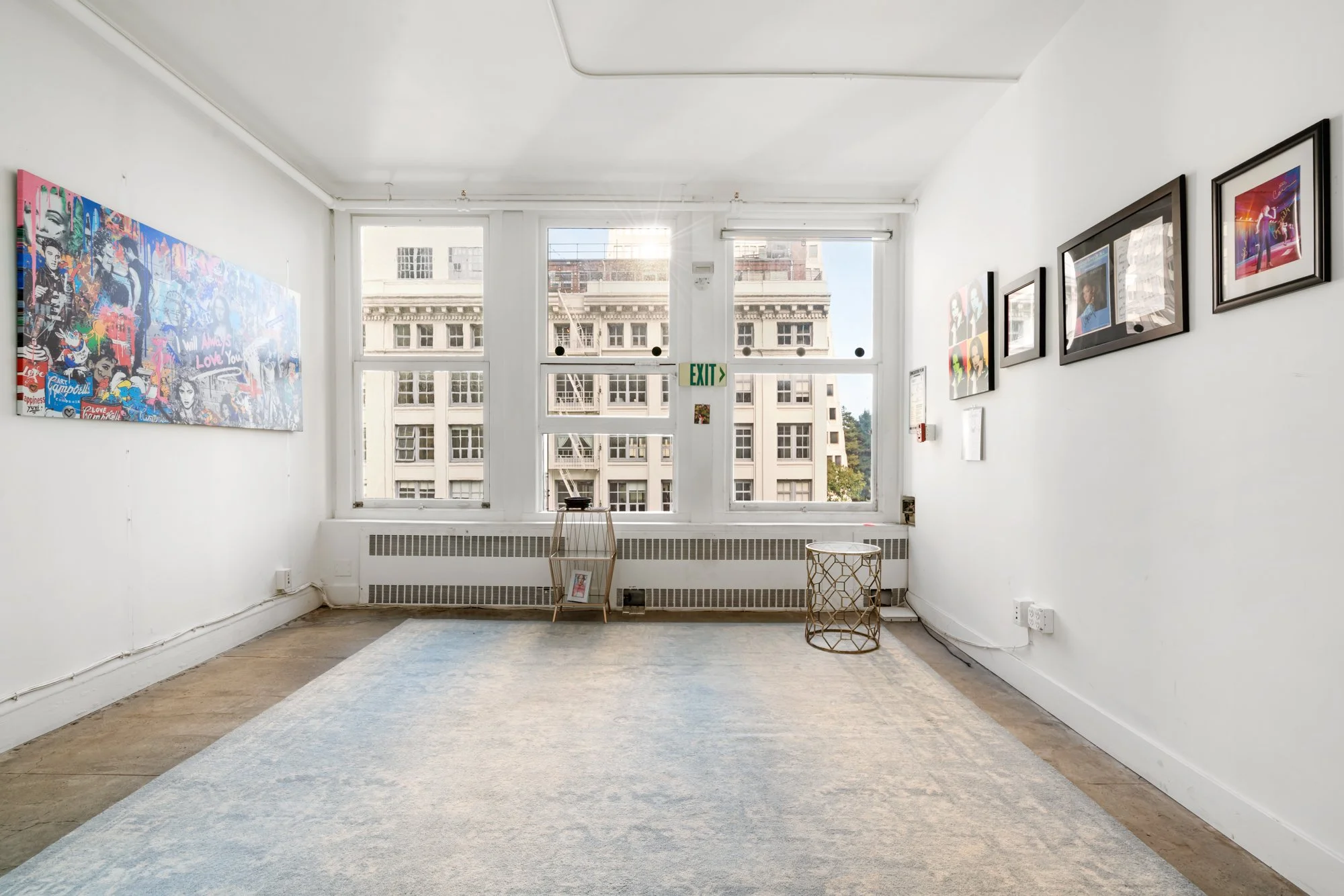 Empty art gallery with framed photographs on white walls, large windows at the back showing city buildings, and a light-colored rug on the wooden floor.