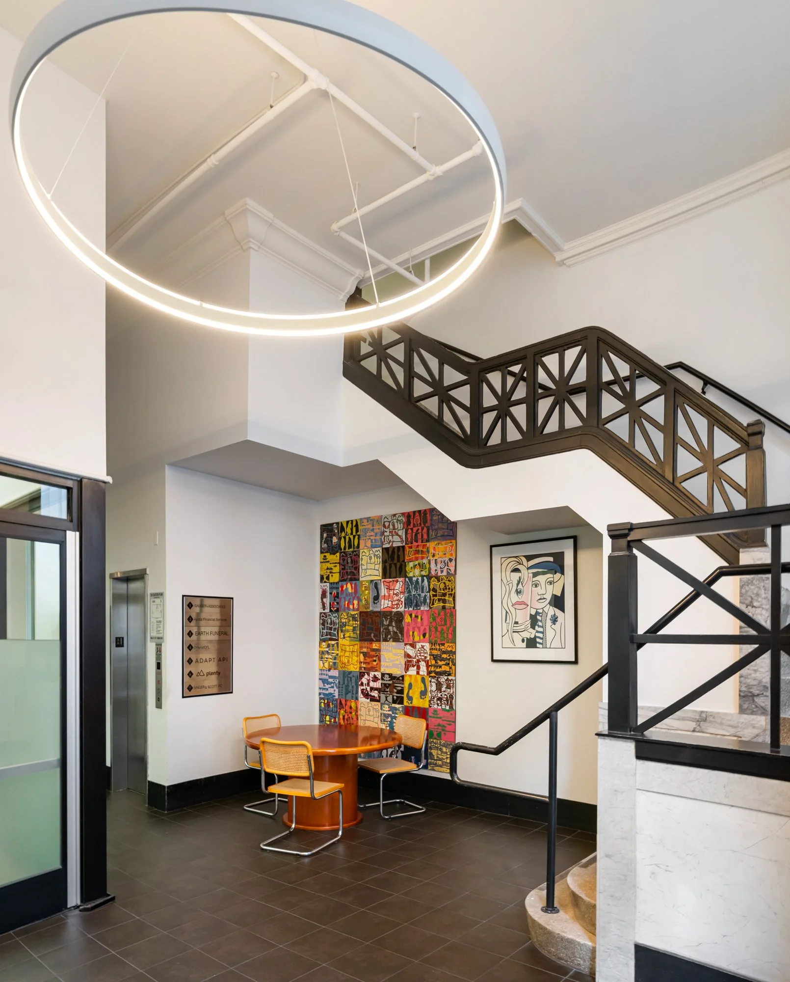 Modern interior lobby with a circular LED ceiling light, a staircase with geometric black railing, a colorful abstract wall art, a framed line drawing, a round wooden table with four orange chairs, and an elevator with a bronze plaque.