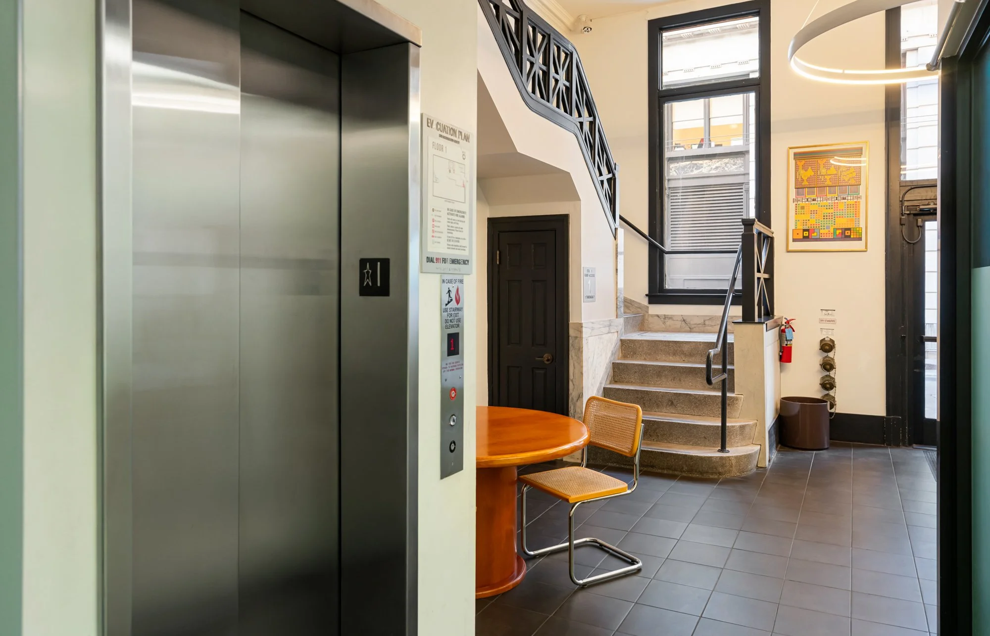 Interior of a building lobby with an elevator, a staircase, and a small round wooden table with a chair. There are large windows, artwork on the wall, a fire extinguisher, and utility controls.