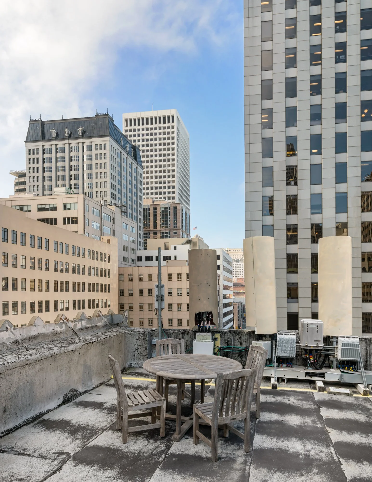 Rooftop patio with a wooden table and chairs, surrounded by tall office buildings on a partly cloudy day.