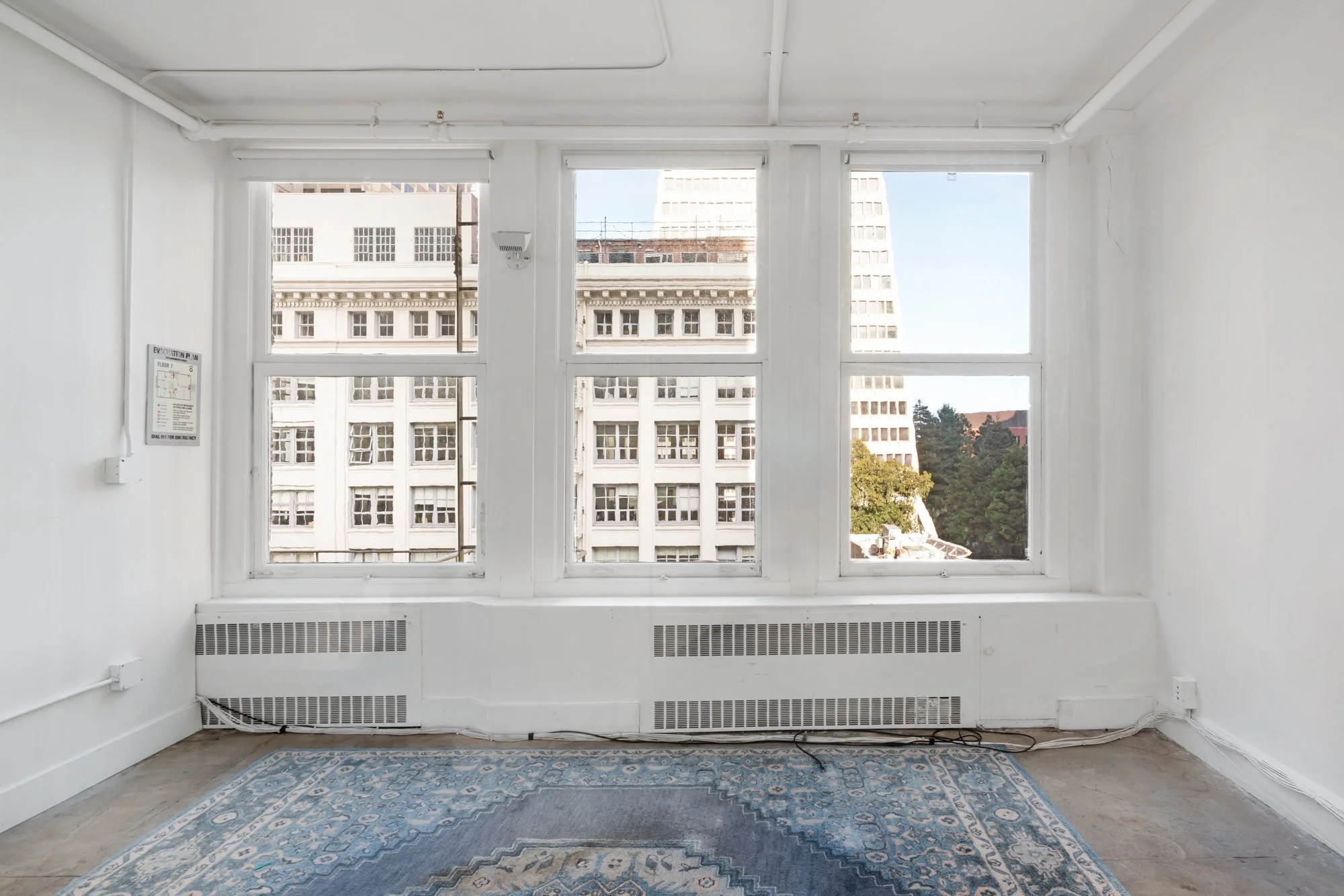 Empty room with large windows showing city buildings and trees, white walls, baseboard heater, electrical outlets, and a patterned rug.