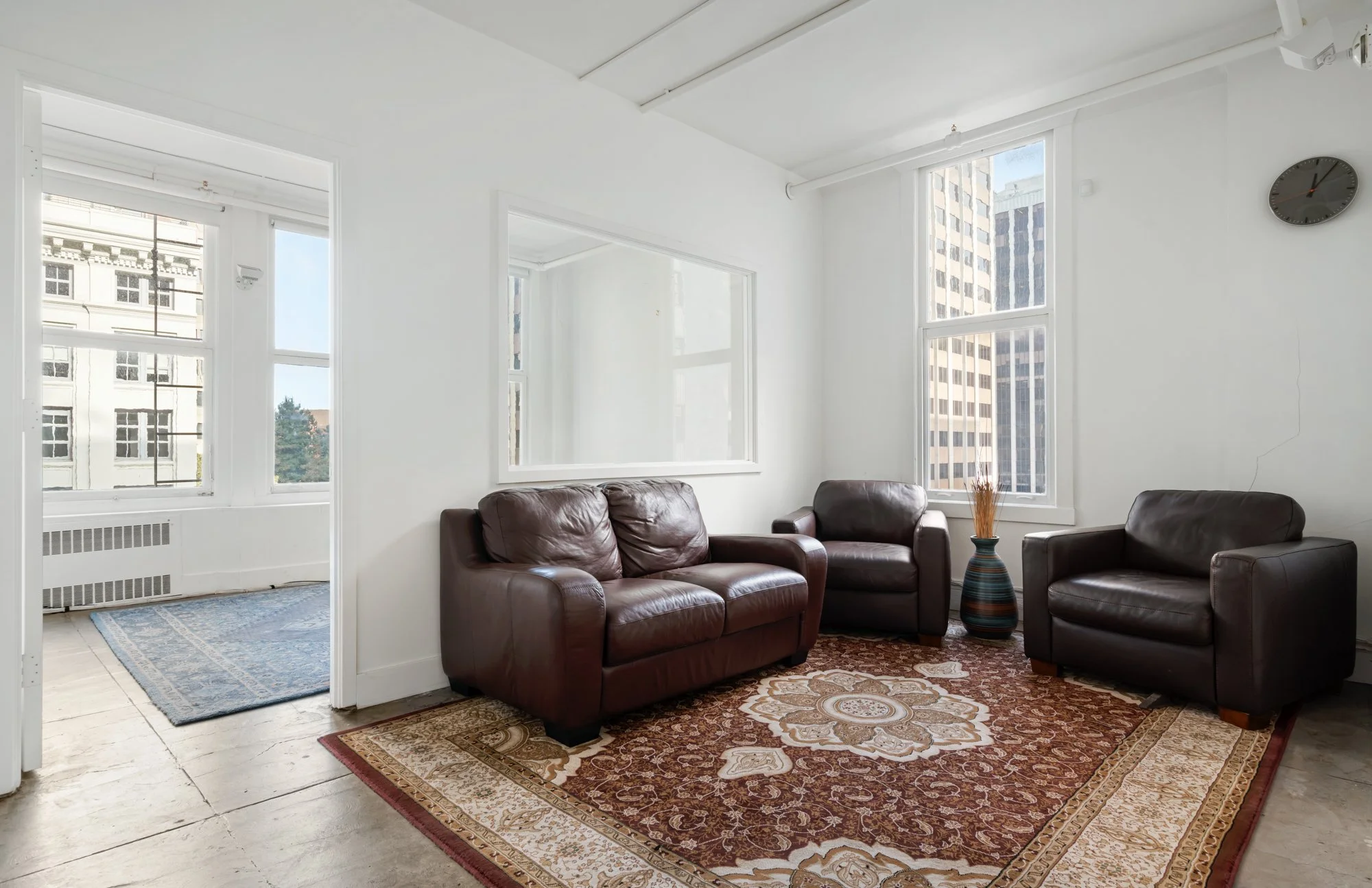 Living room with white walls, large windows with city views, a brown leather sofa and two matching armchairs, a decorative rug, a tall vase with dried plants, and a round wall clock.