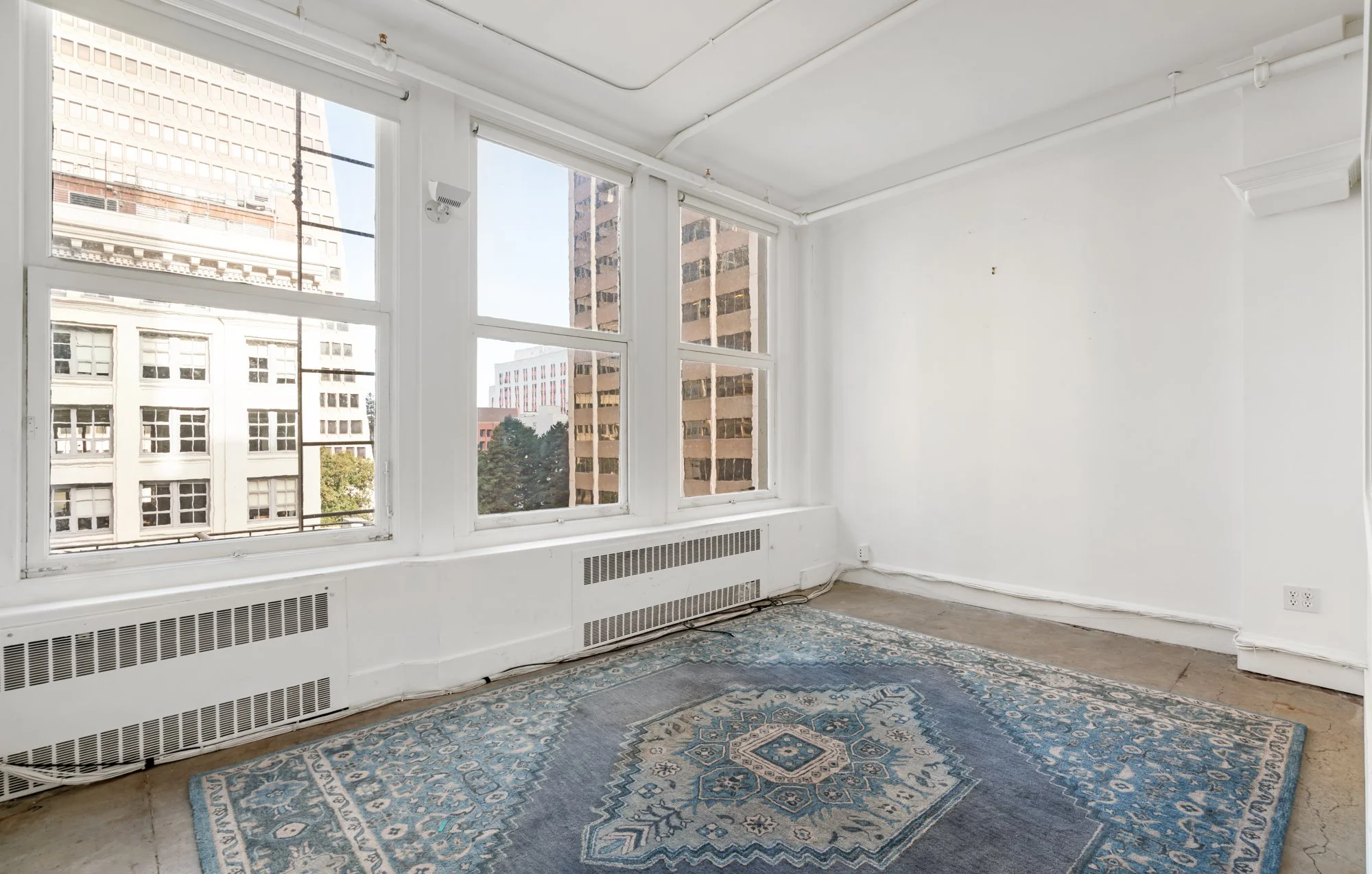 Empty room with large windows showing city buildings, white walls, radiator, and a blue patterned rug on the floor.