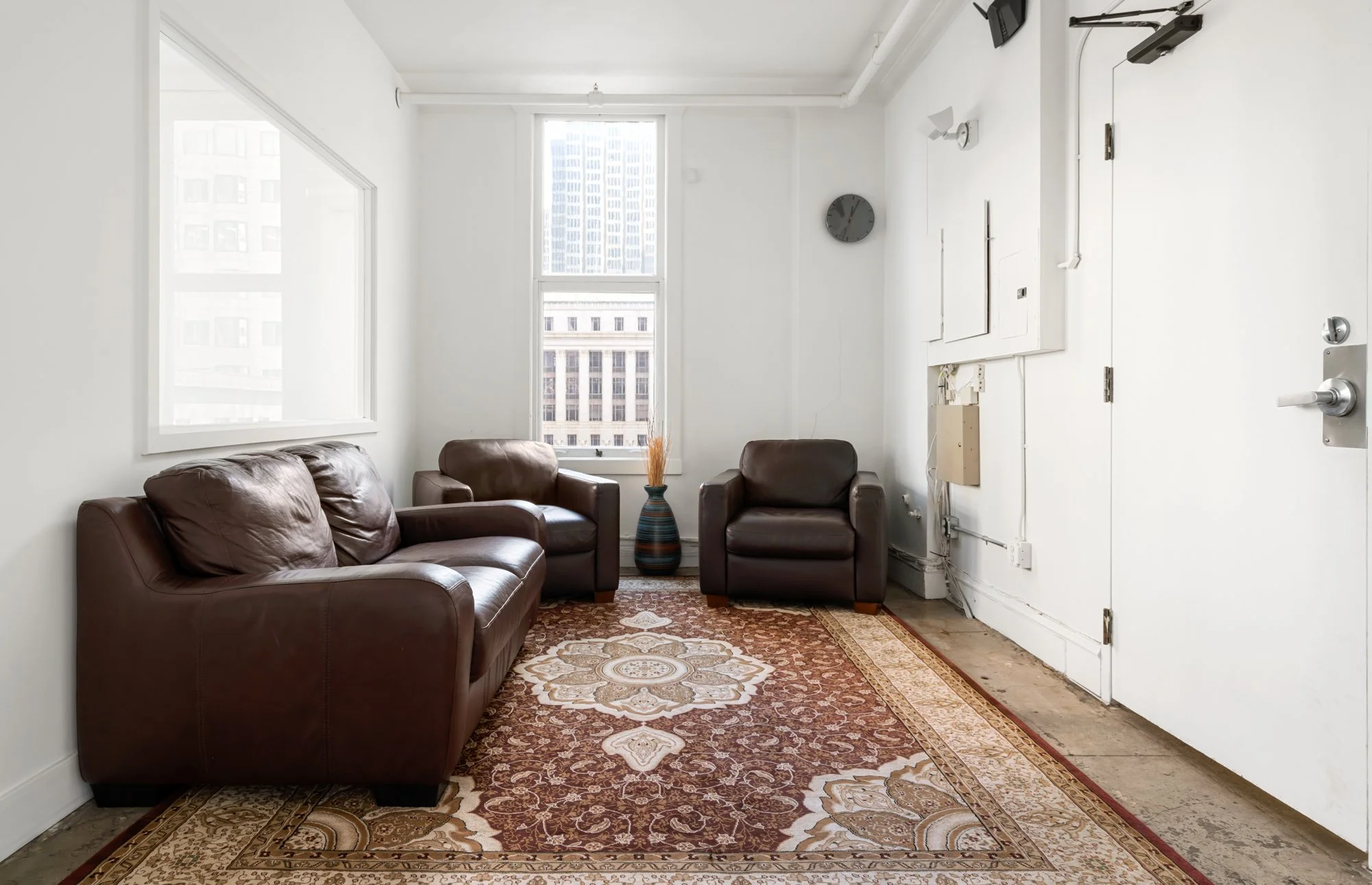 Living room with three brown leather armchairs, a colorful vase with dried plants, a patterned area rug, white walls, a large window with a city view, a wall clock, and electrical panels.