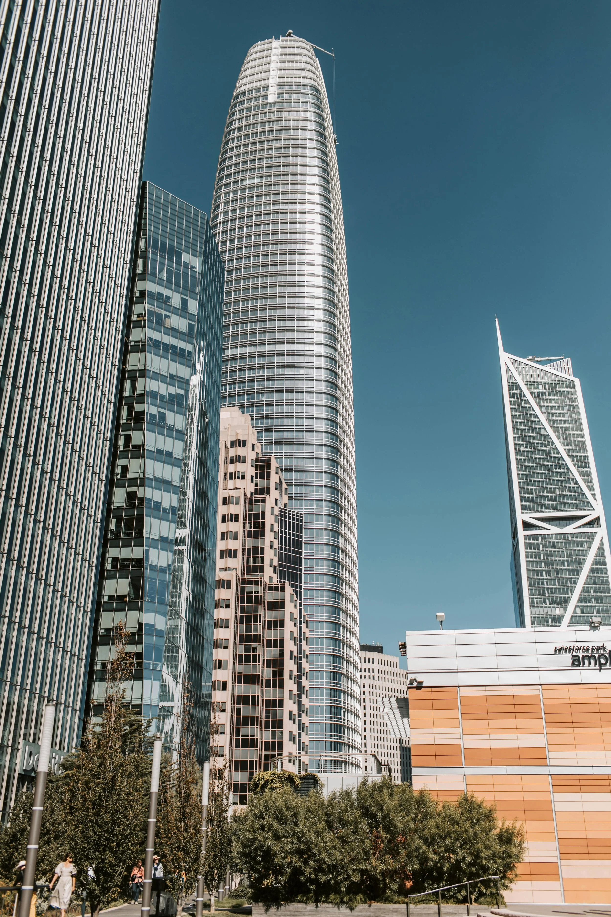 Tall modern skyscrapers in a city, with a clear blue sky and greenery at the base.